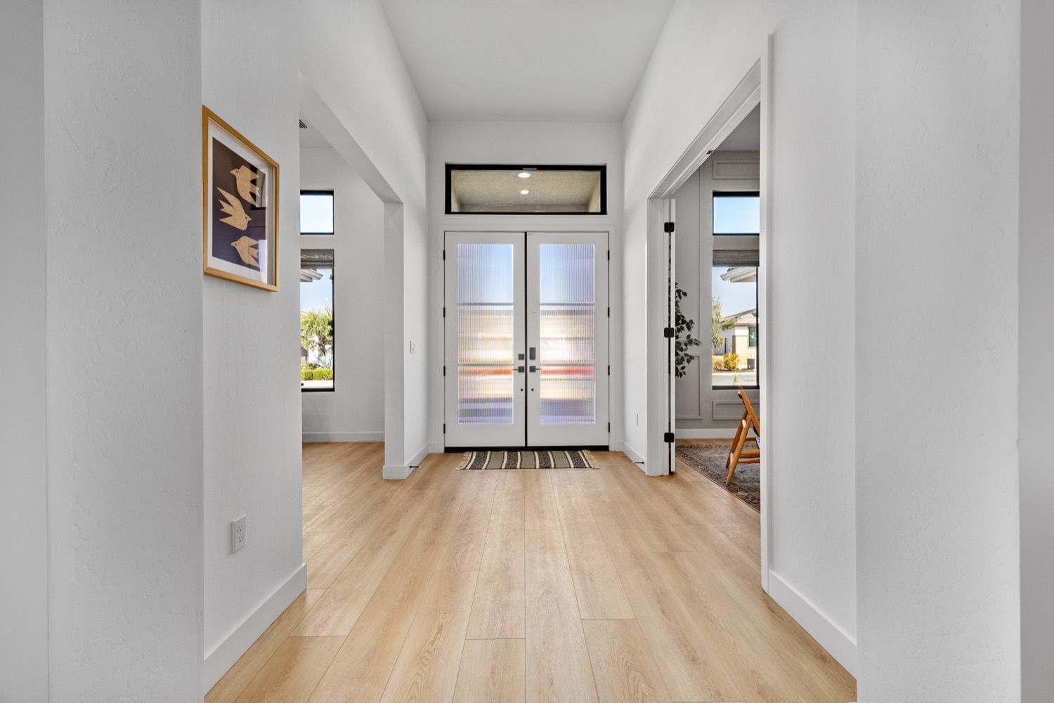 Hallway with light wood floors, white walls, and double glass doors leading outside.