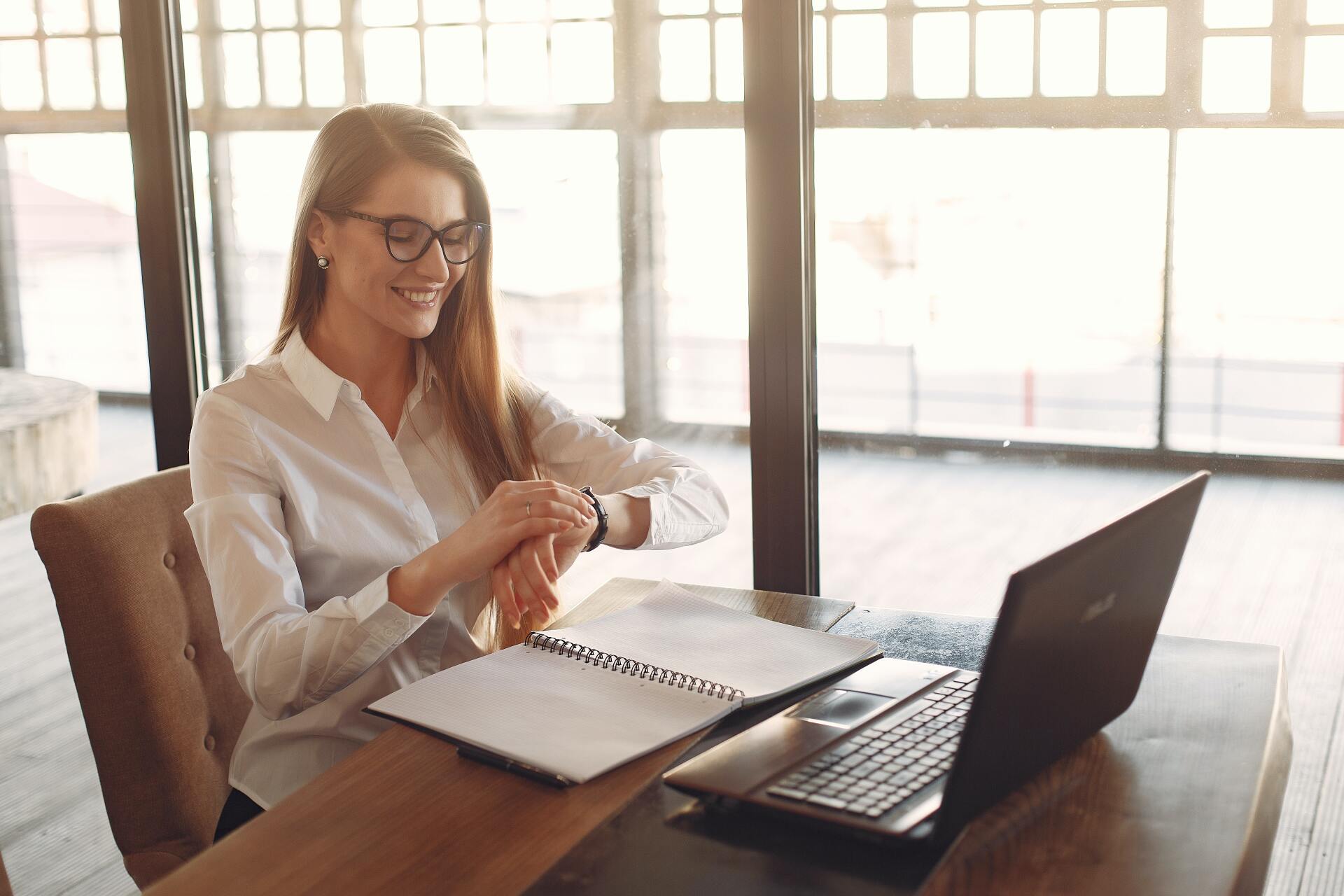 A woman is sitting at a desk with a laptop and looking at her watch.