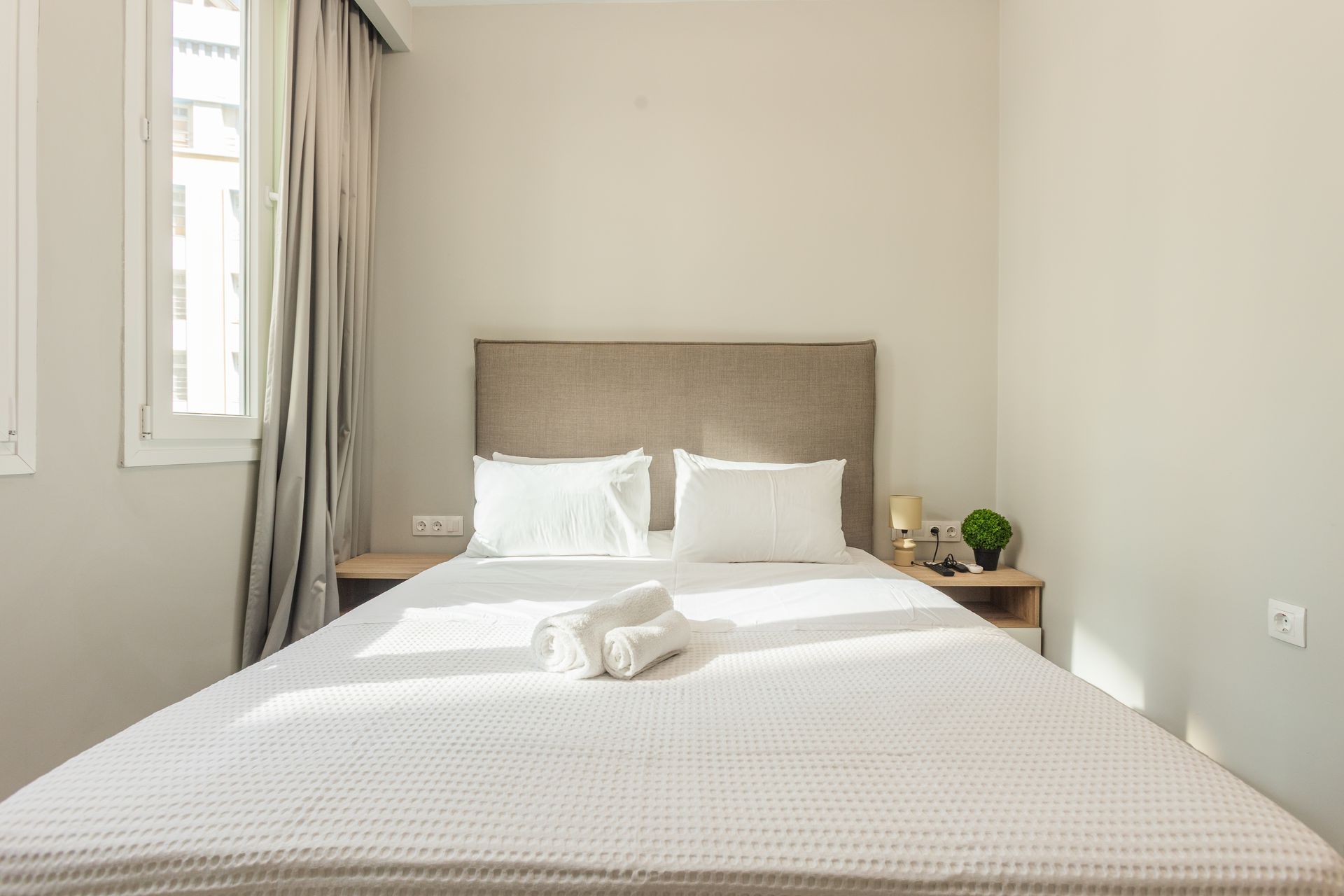 Bedroom with a bed, two pillows, folded towels, and two nightstands. Window with curtains on the left.