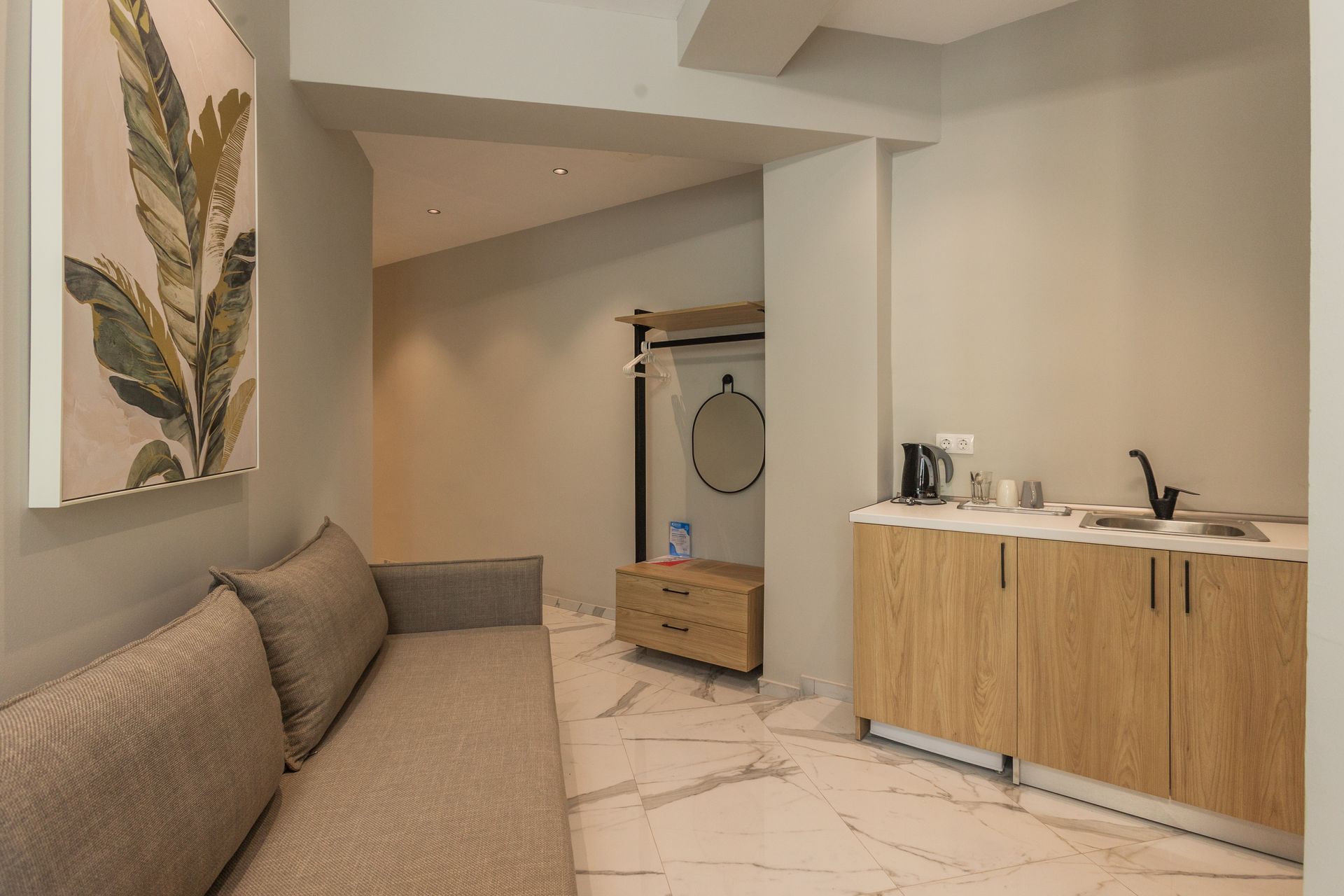 A neutral-toned hotel room interior with a sofa, kitchenette, artwork, and a hanging clothes rack.