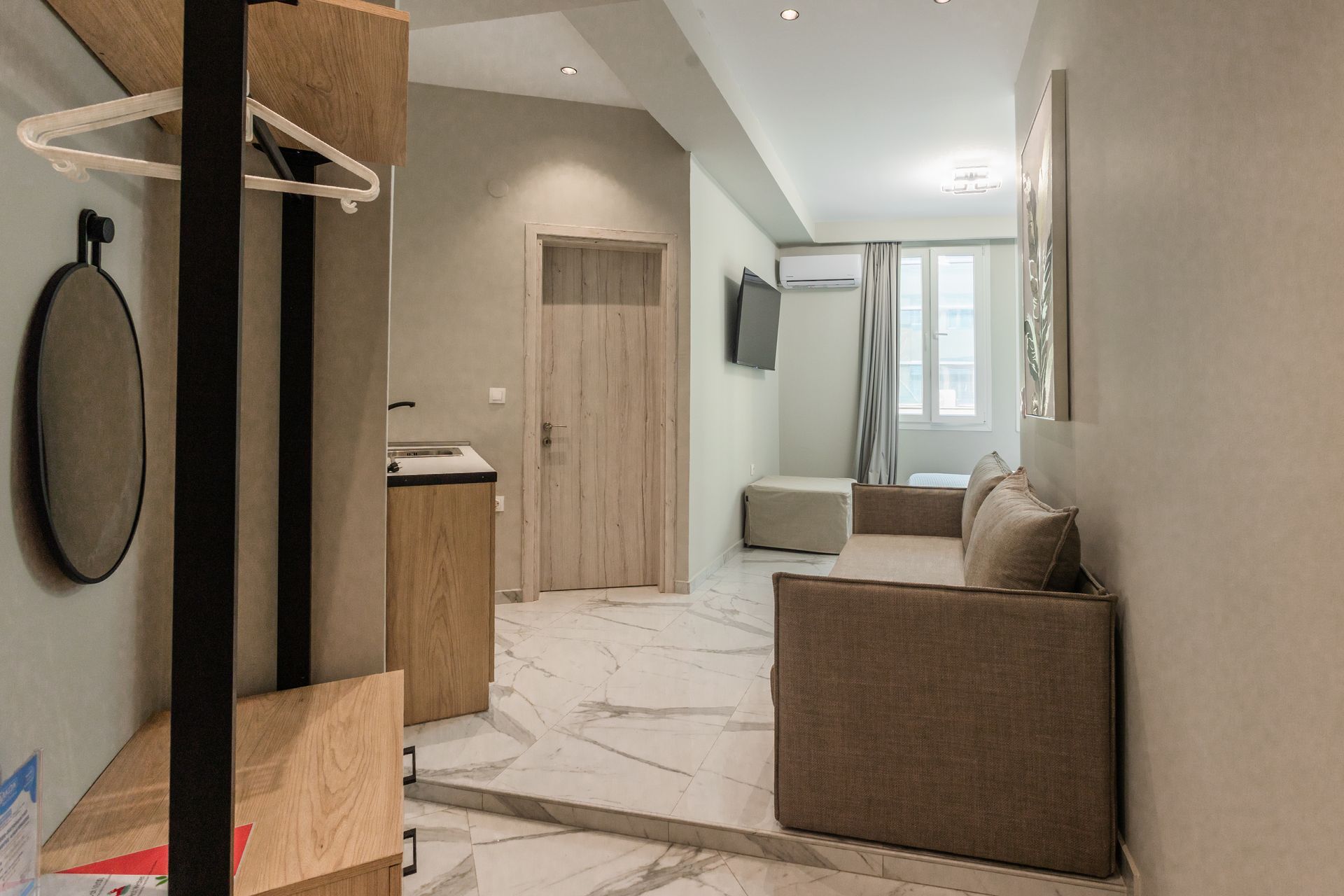 Small apartment interior with sofa, TV, door, mirror, and hanging rack. Marble-look floor.