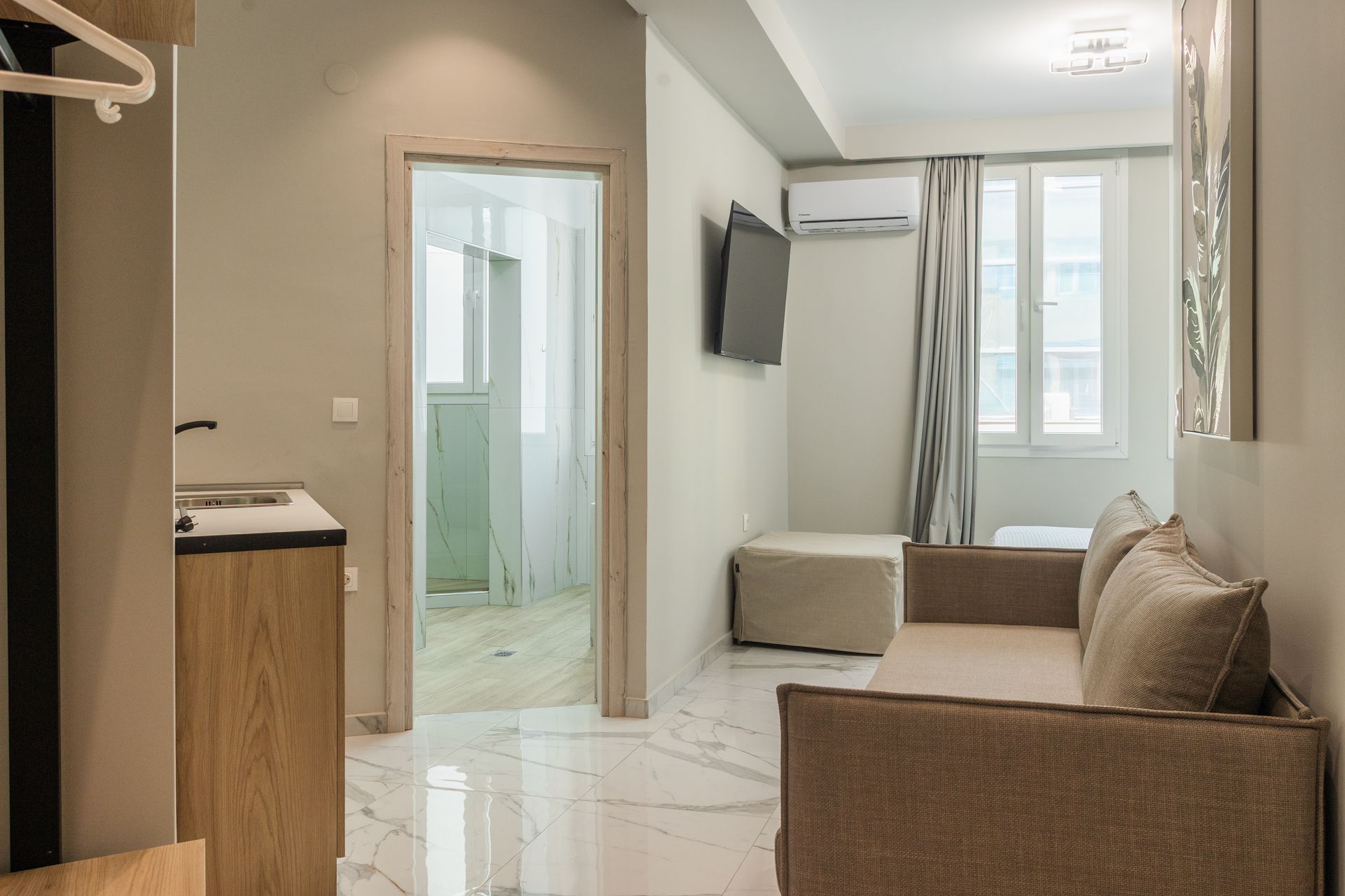 A small apartment with a sofa, TV, and open doorway leading to a balcony. Beige and white tones.