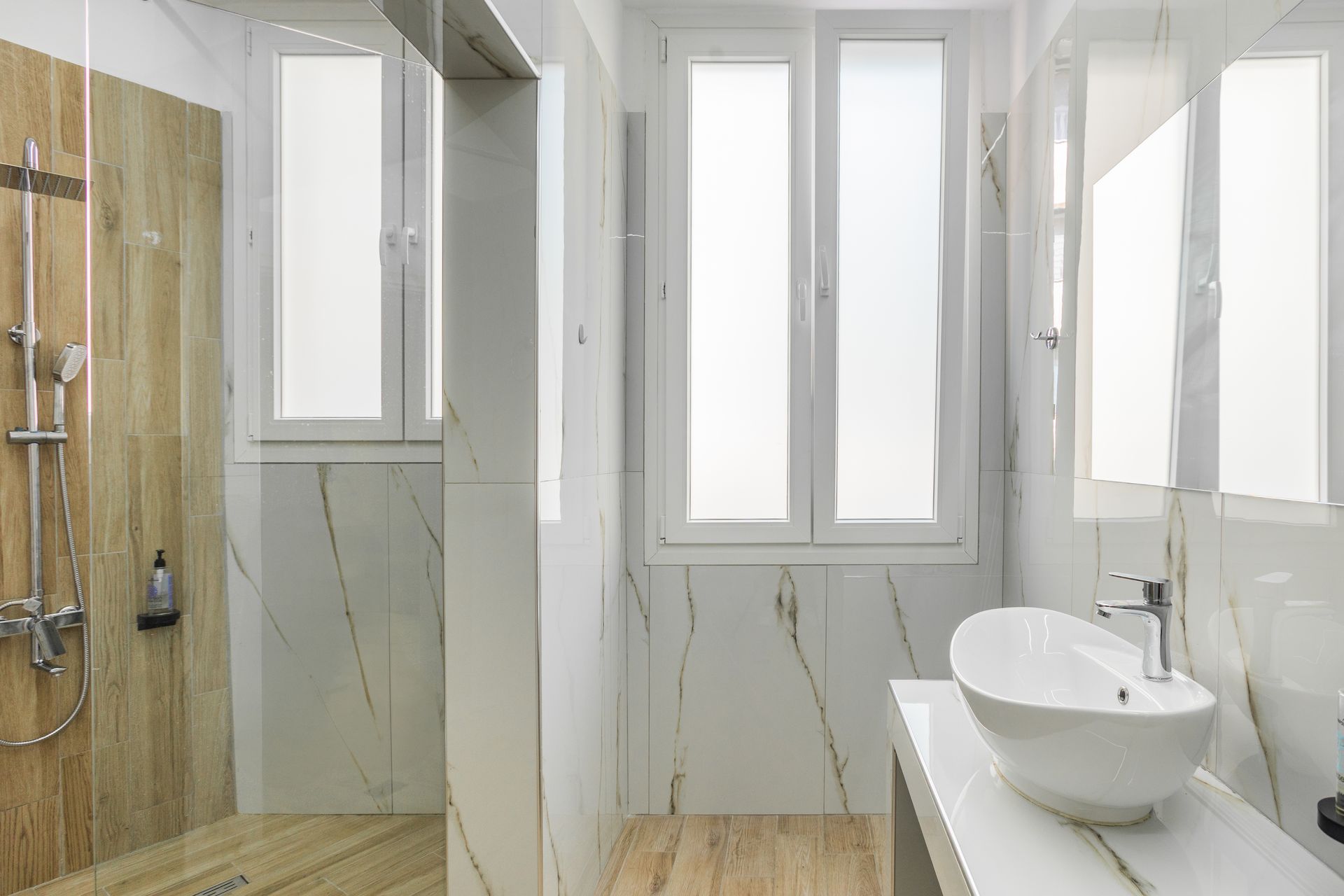 Bathroom with white marble walls, wood-look shower, and a white, oval-shaped sink. Windows provide natural light.