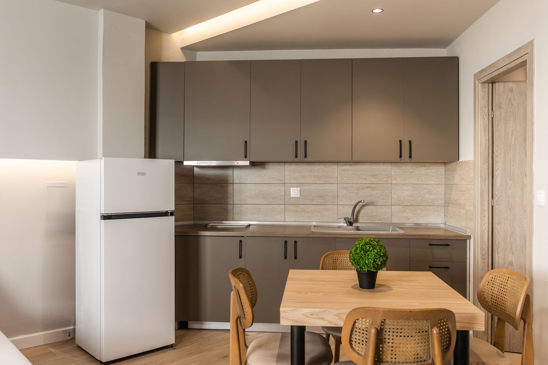 A small kitchen with gray cabinets, a fridge, a dining table with chairs, and a small plant.