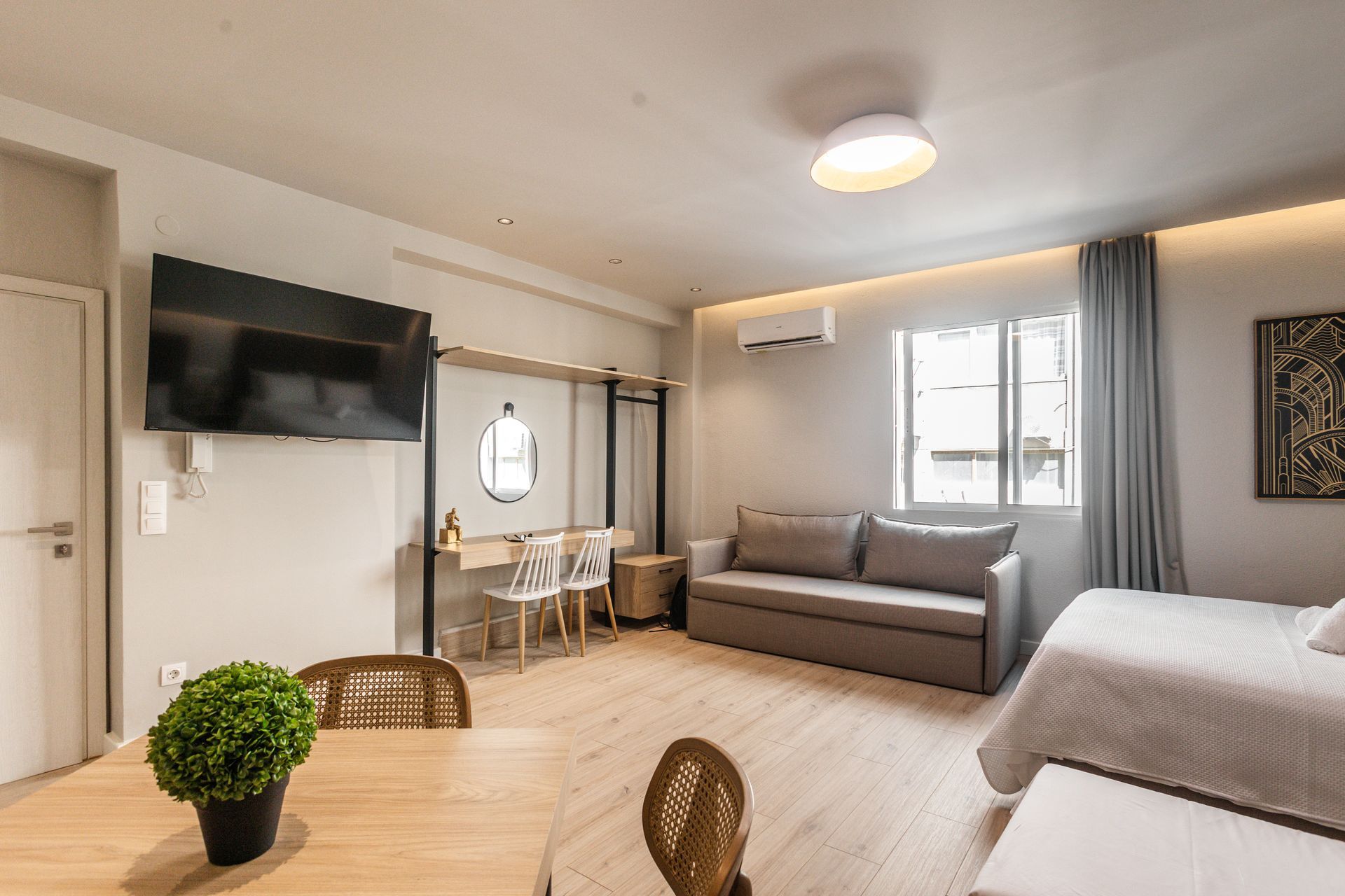 Studio apartment with light wood floors, sofa, bed, desk, and TV.