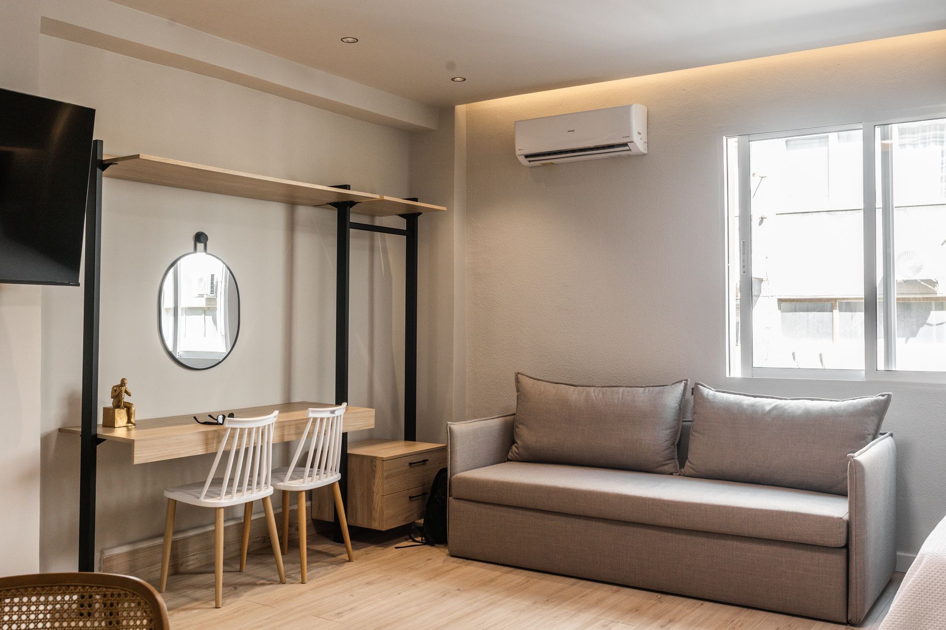 Modern hotel room with sofa, desk, window, and air conditioner on a beige wall.