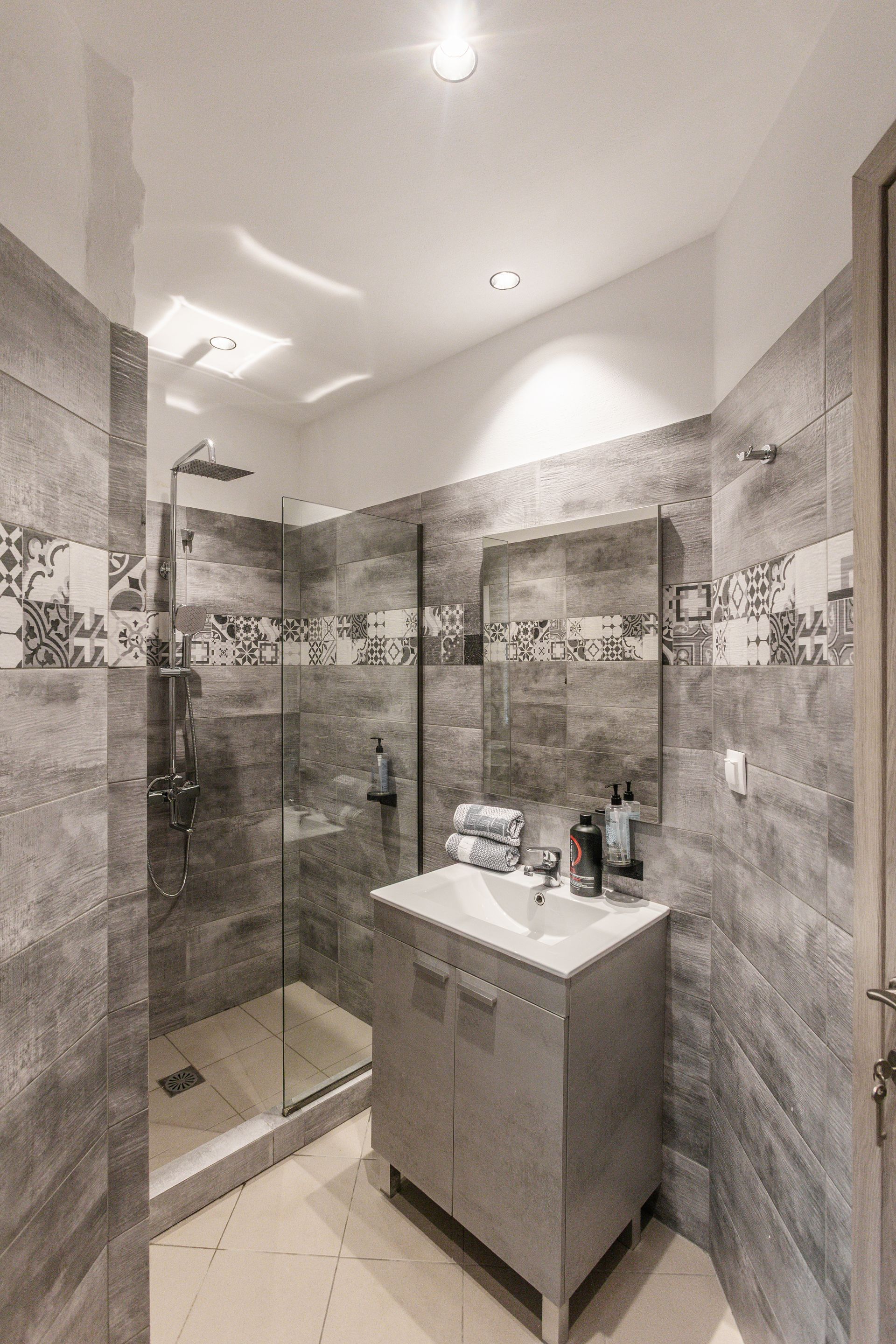 Small, modern bathroom with gray tiled walls and glass shower. A vanity and mirror are centered.