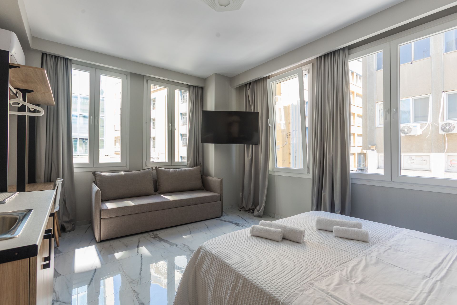 Bright studio apartment with large windows, sofa, bed, and TV.