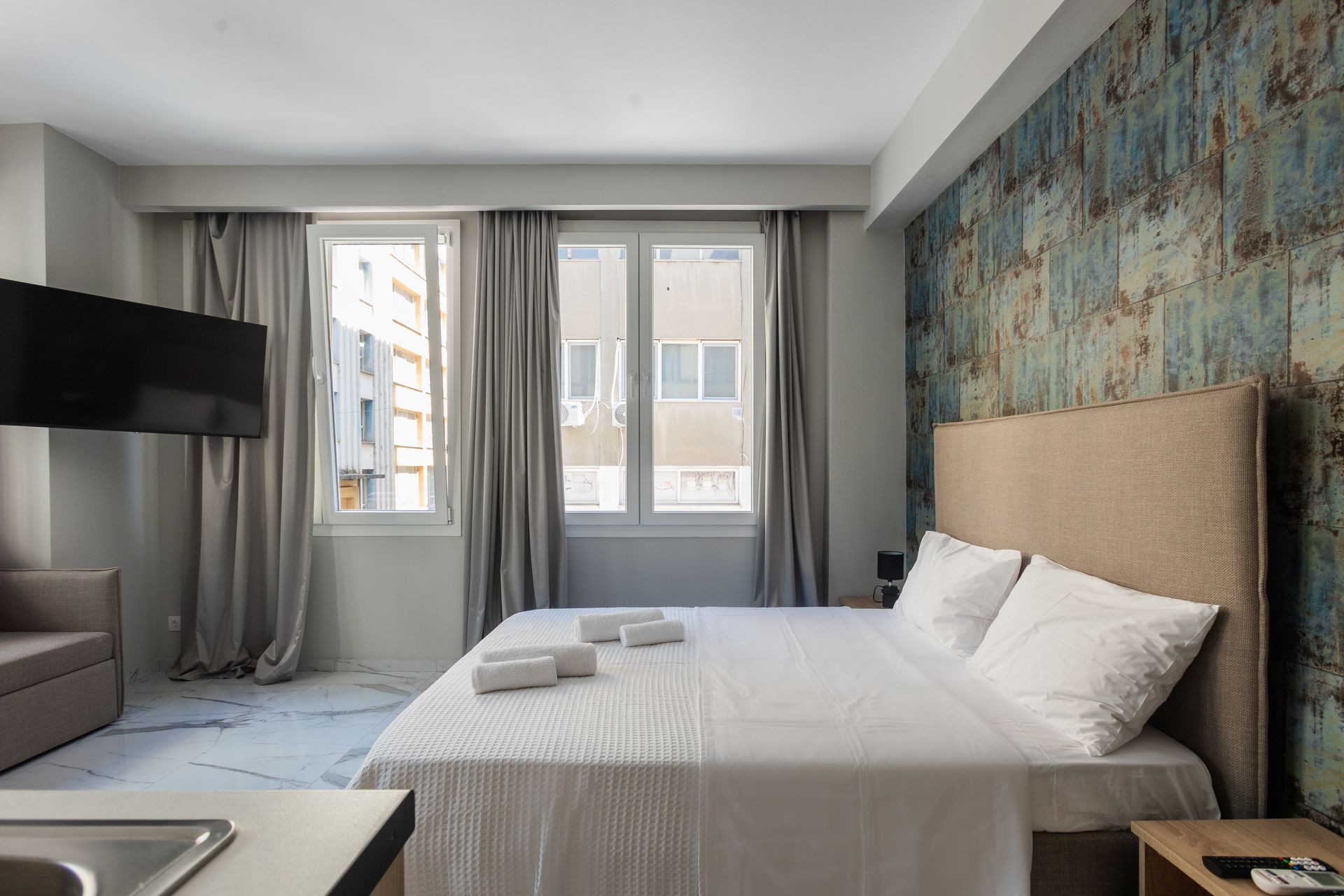 Bedroom with a large bed, TV, sofa, and window with grey curtains. Textured wall behind bed.