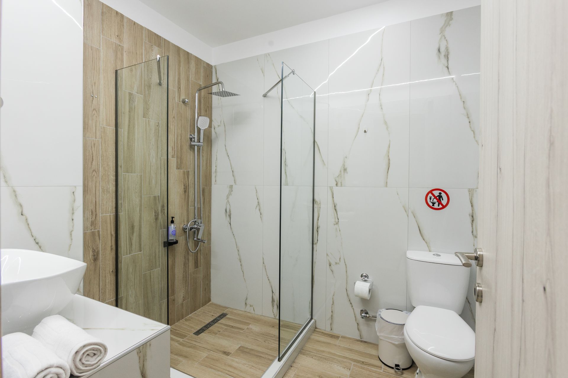 Modern bathroom with white and marble tile, glass shower, toilet, and rolled towels.