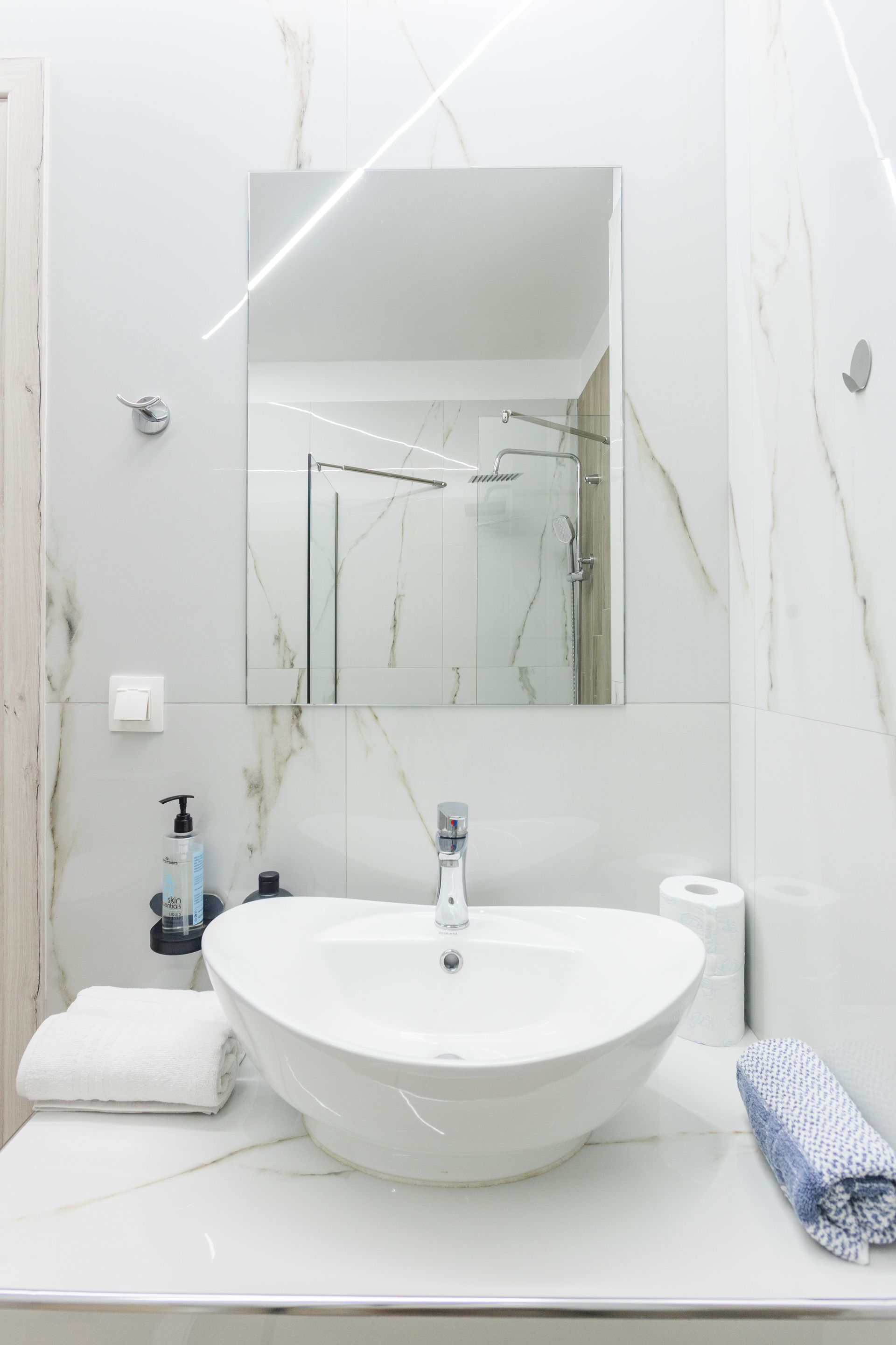 White bathroom with a vessel sink, mirror, and marble-like tile. Towels and toiletries are visible.