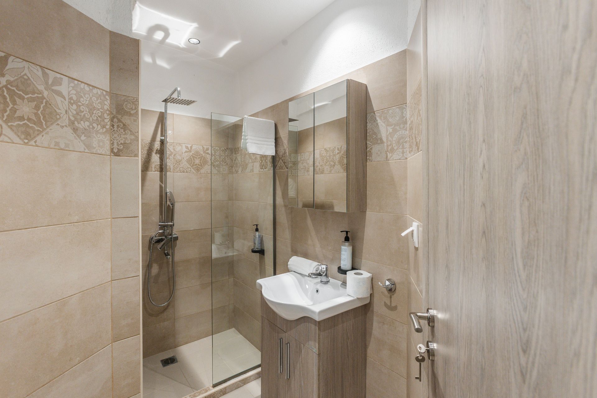 Small bathroom with shower, sink, and cabinet; beige tiles and light wooden door.