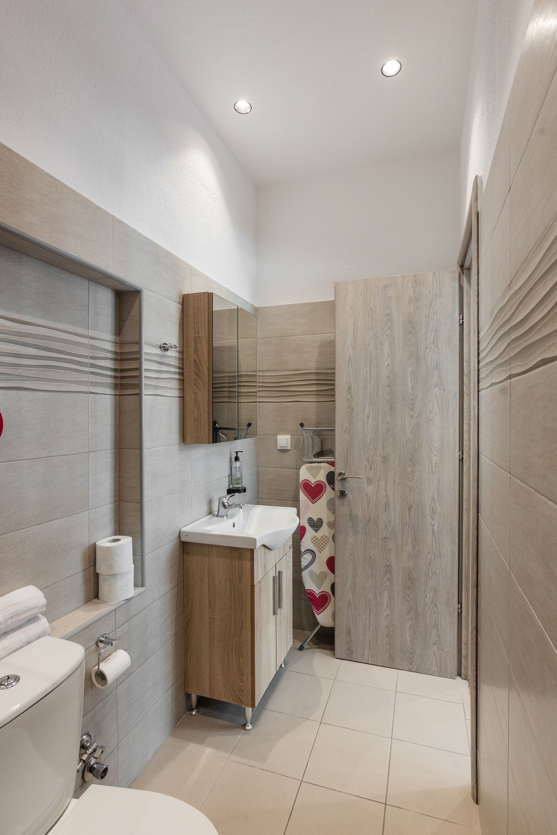 A small, modern bathroom with neutral-toned tiles, a toilet, sink, and closed wooden door.