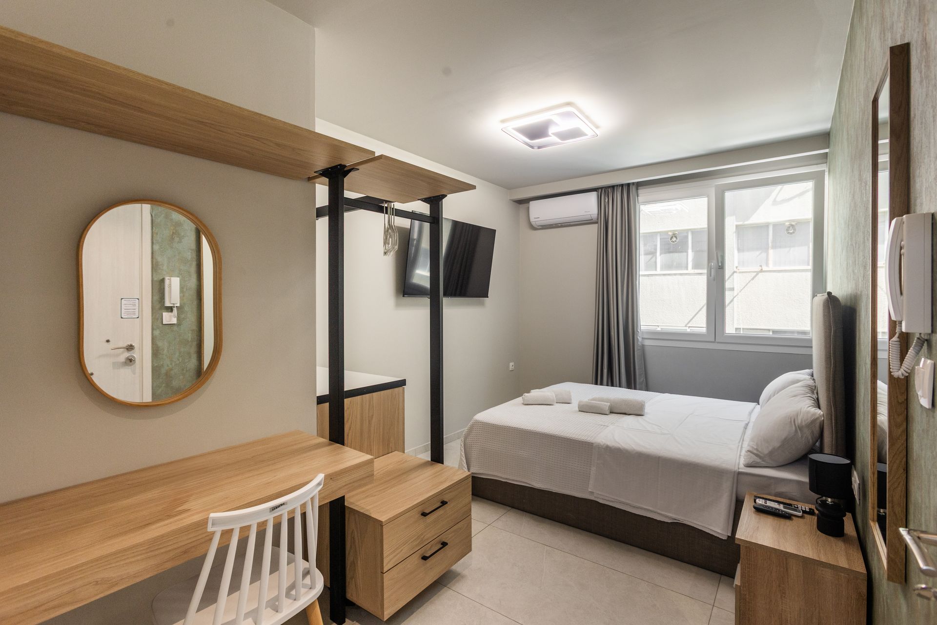 Small bedroom with a bed, desk, storage, and a mirror. Grey and wood tones, window with a curtain.