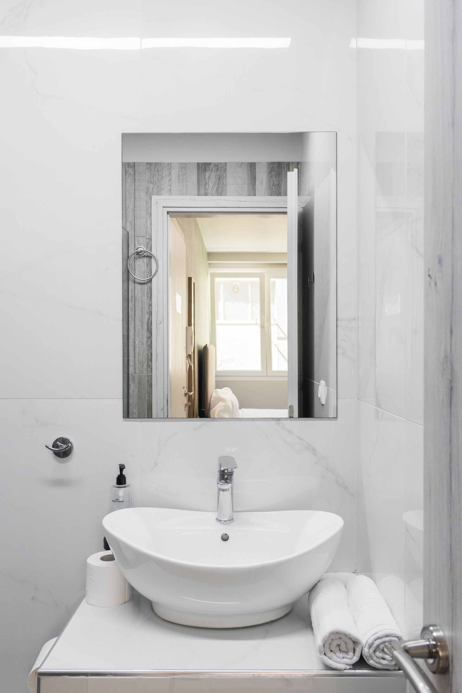 White modern bathroom with a sink, mirror, and towels, reflecting a glimpse of a bedroom.