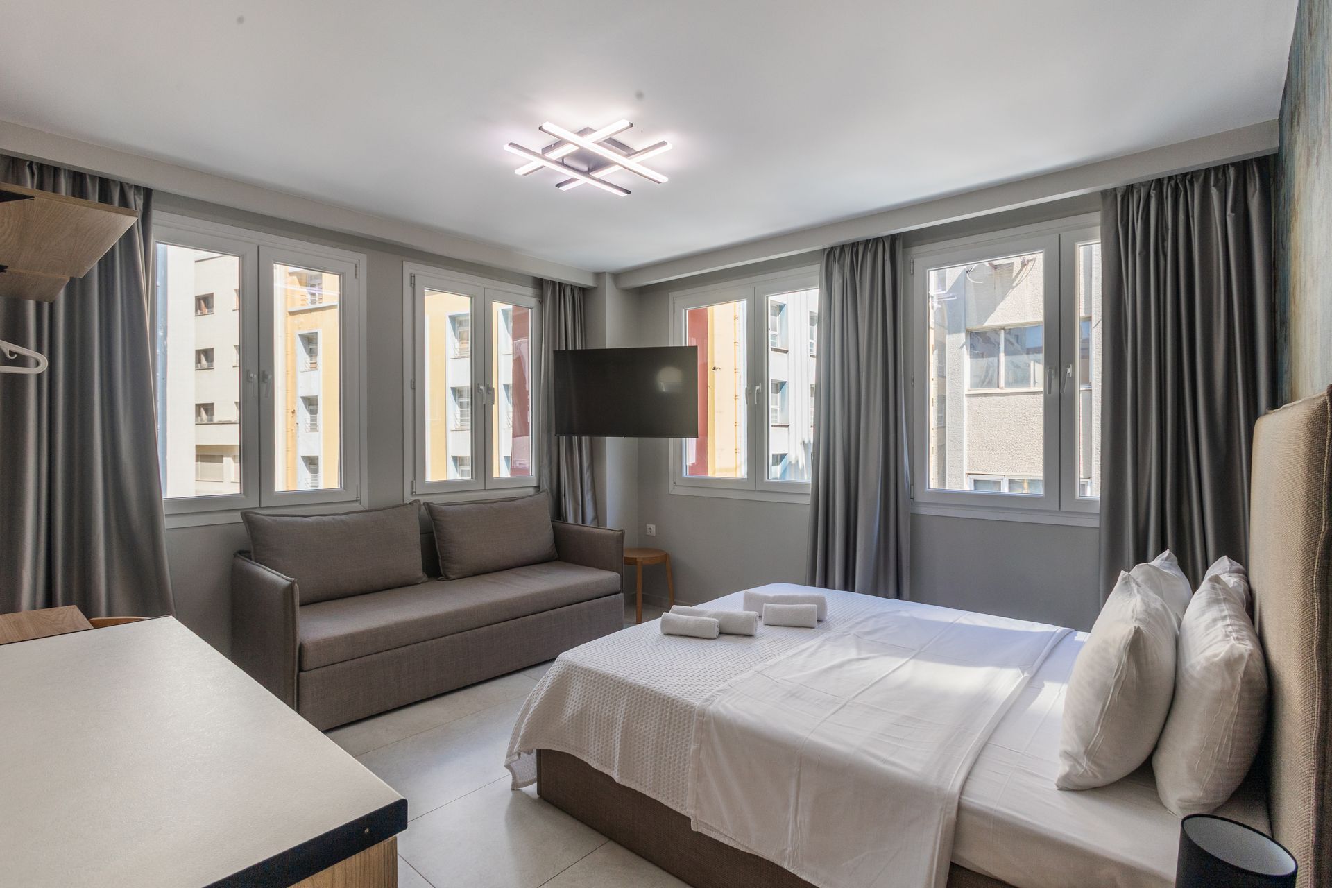 Bedroom with bed, sofa, windows with grey curtains, and TV.
