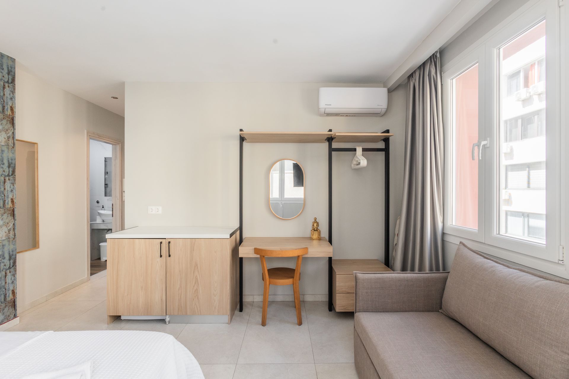 Interior of a hotel room with a desk, mirror, closet rack, and couch near a window.