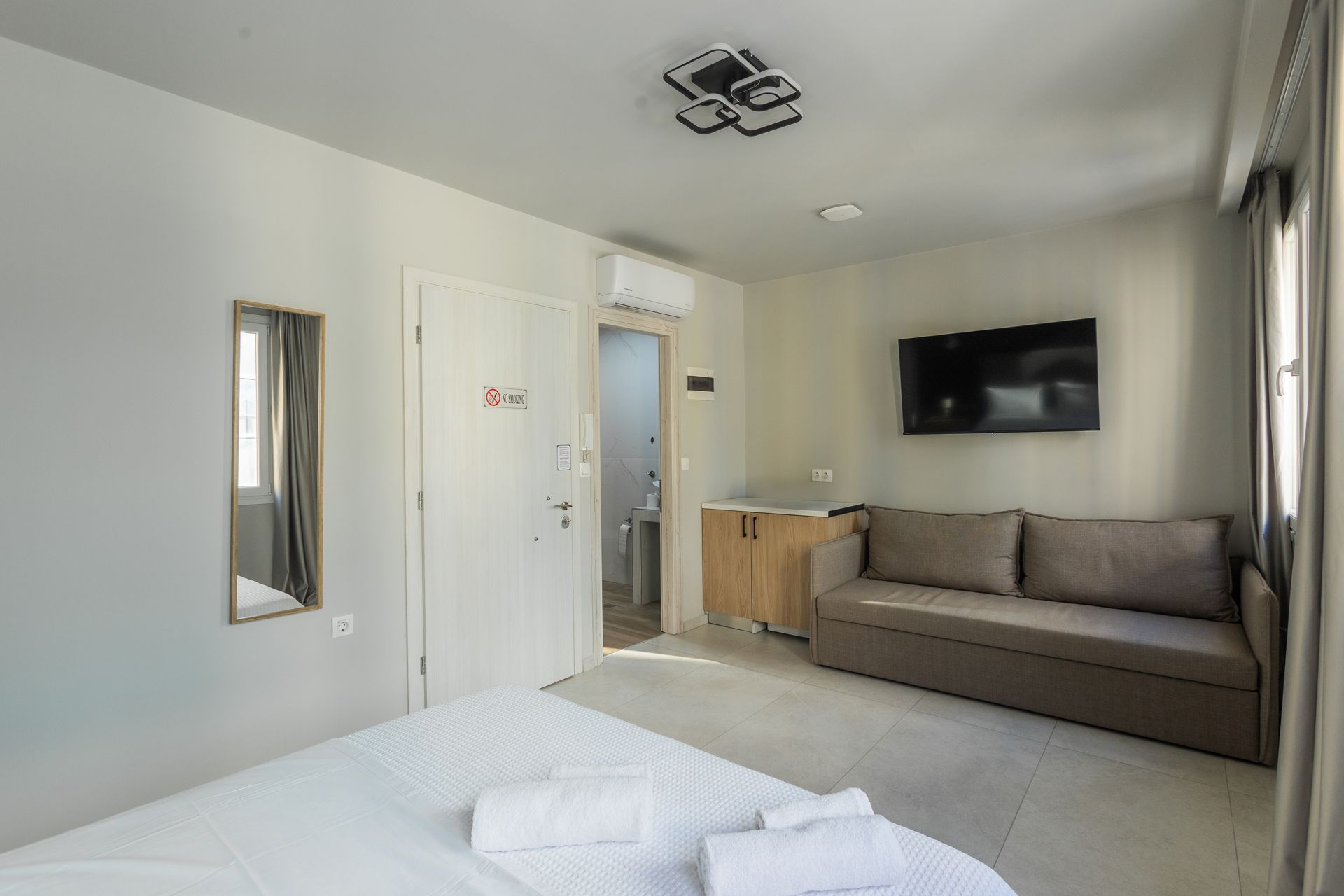 Bedroom with white walls, bed, sofa, TV, and open door to a small bathroom.