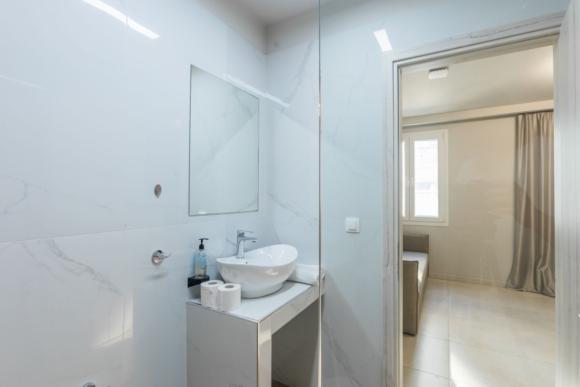White modern bathroom with a sink, mirror, and doorway to a room with a window and a couch.