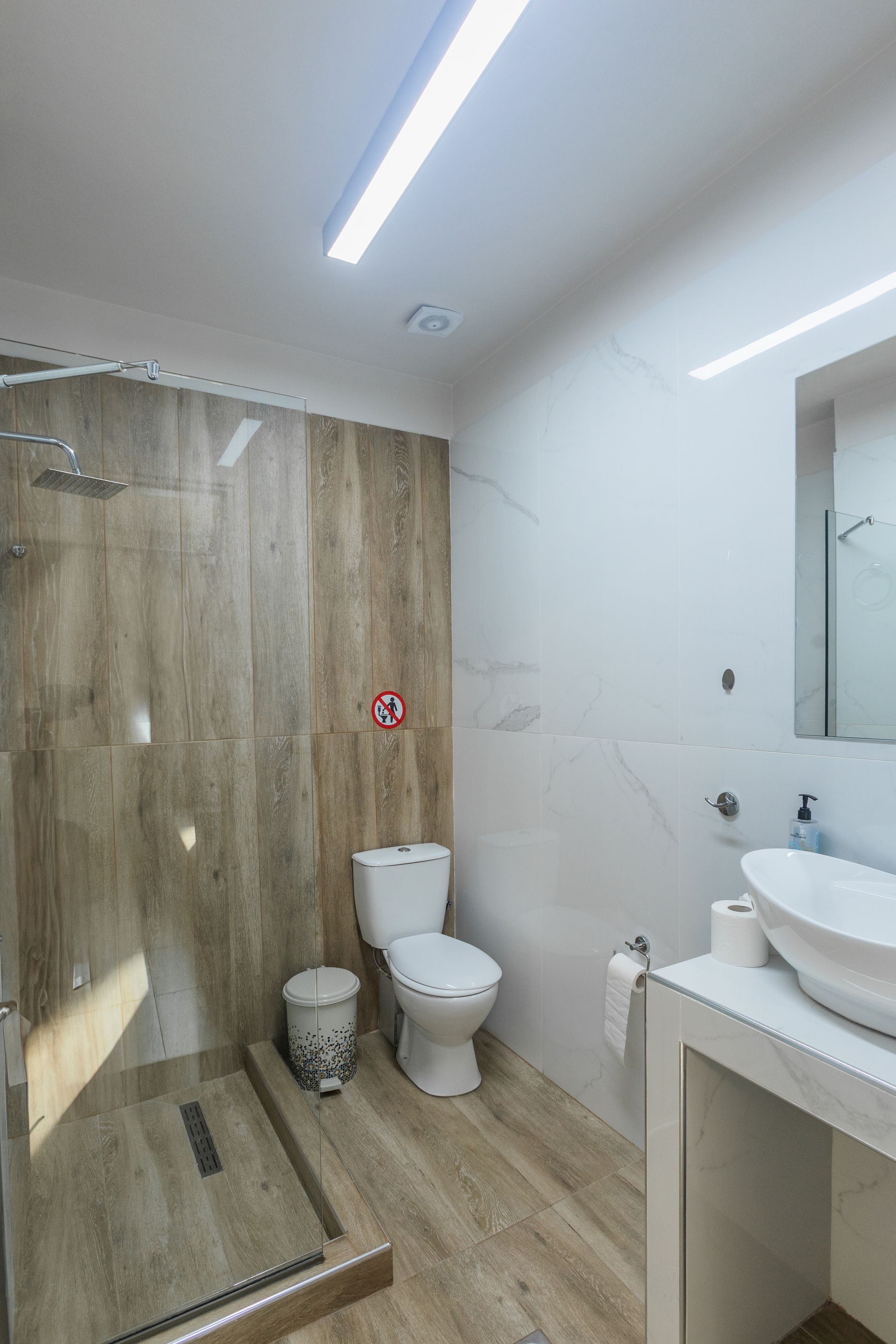 Bathroom with a walk-in shower, toilet, and a sink on a white countertop. Neutral-toned tile and wood-look flooring.