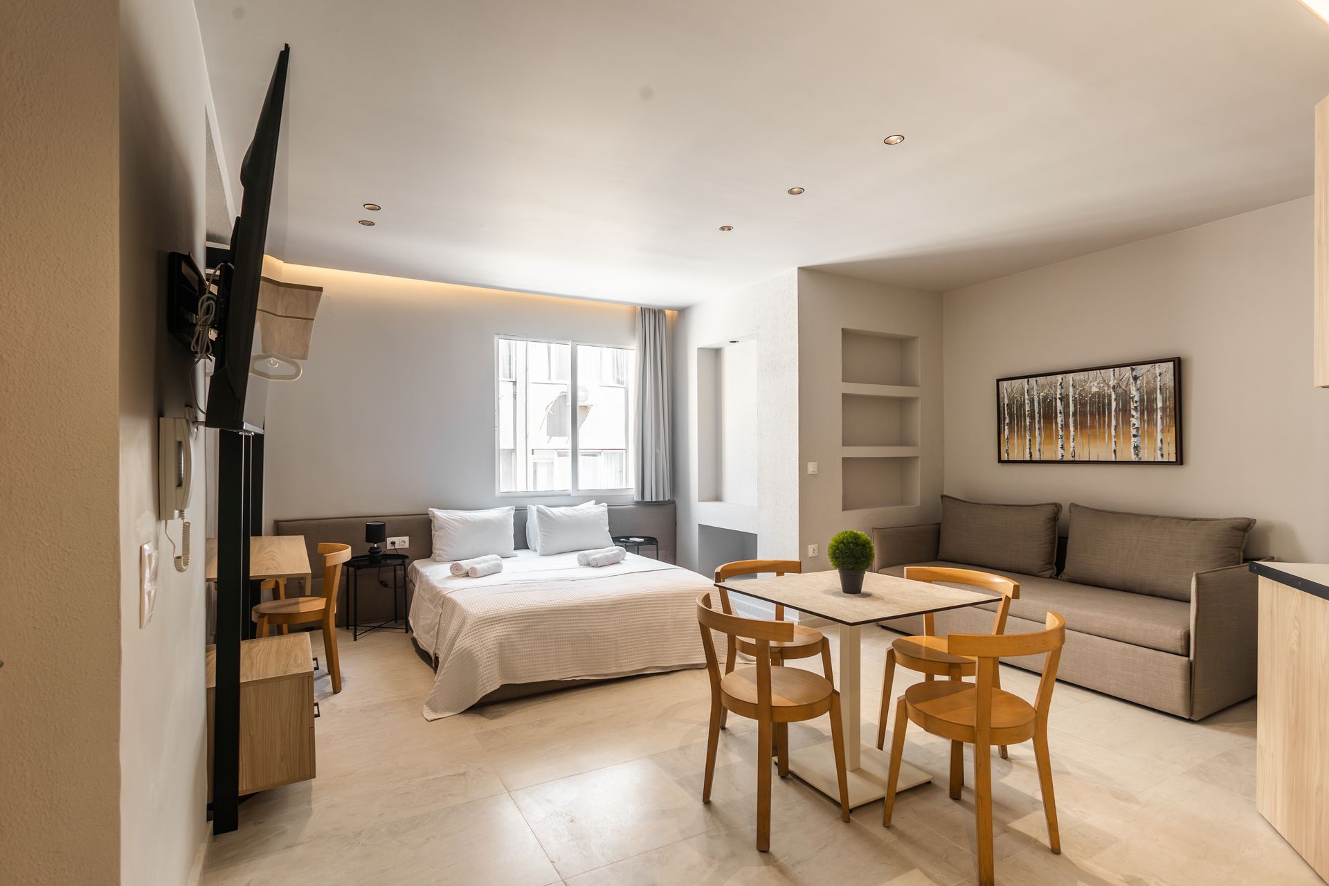 Studio apartment interior with a bed, couch, table, chairs, and a mounted TV.