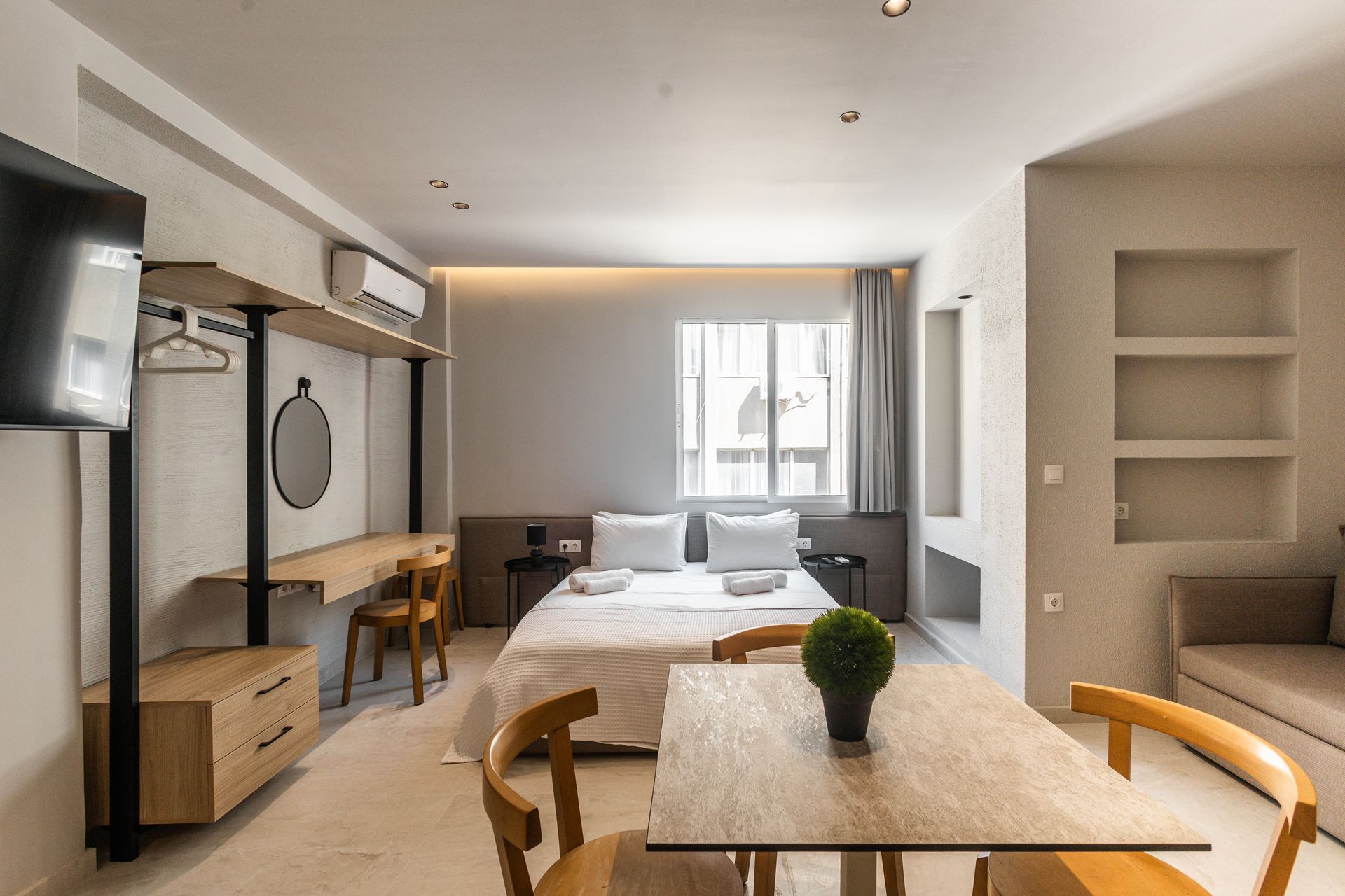 Modern hotel room: bed, desk, table, seating. Neutral colors, natural wood accents.