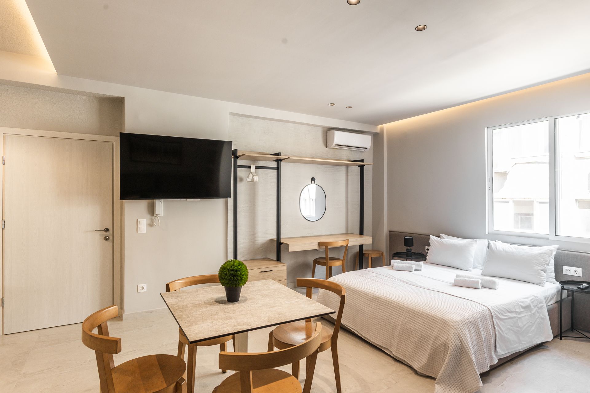 Modern studio apartment with bed, table, chairs, TV, and open closet. Bright, neutral colors.