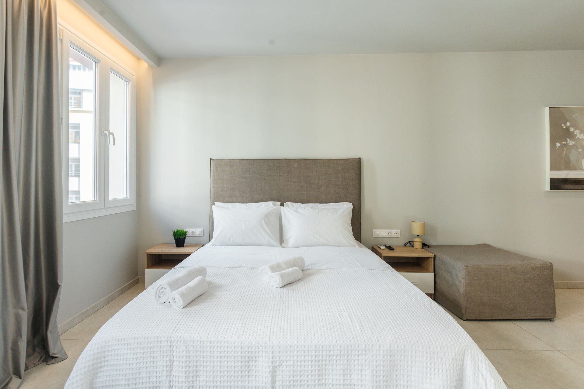 Bedroom with a bed, nightstands, window, and a small ottoman. Neutral colors, natural light.