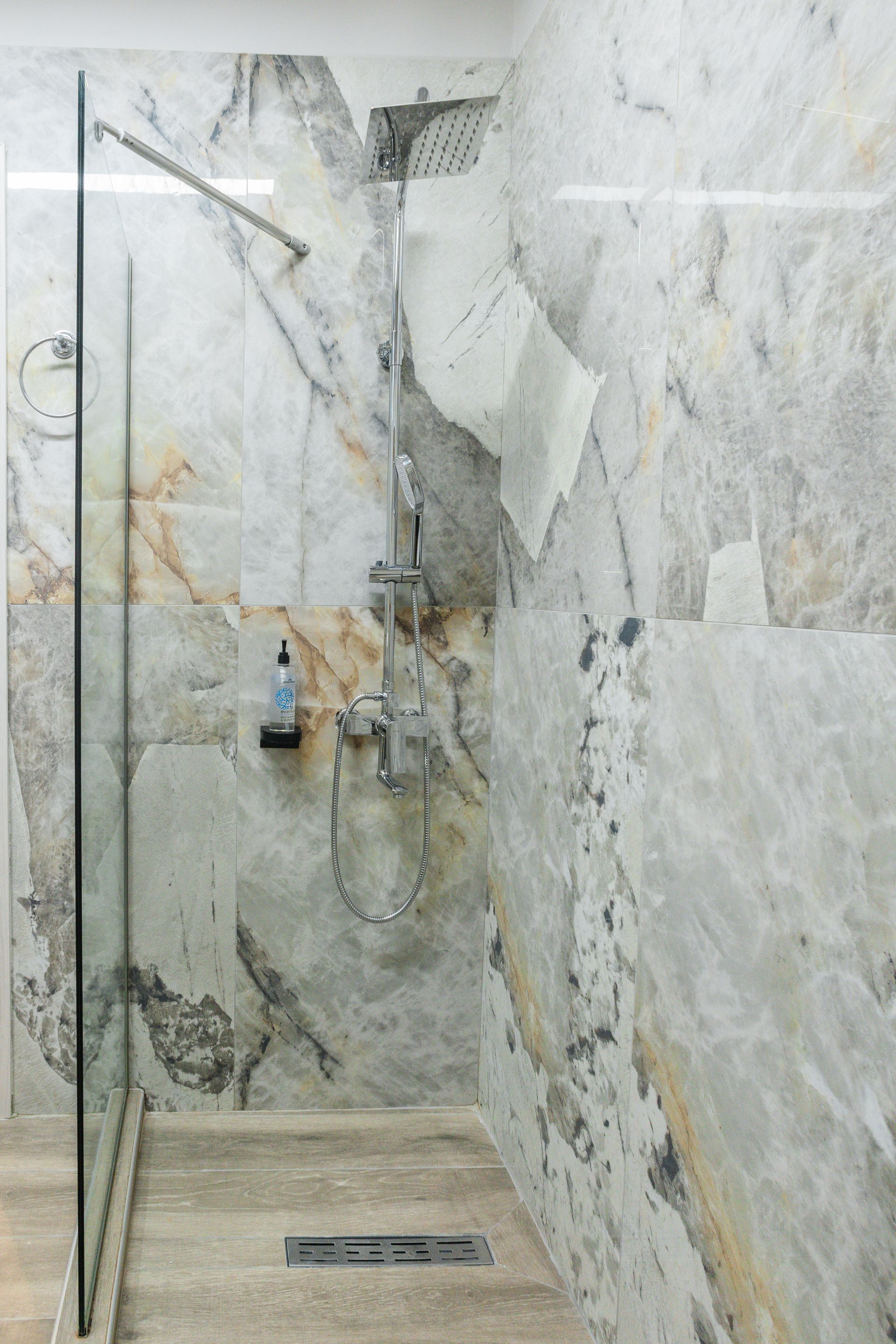 Shower with marbled walls and glass door; handheld showerhead, soap dispenser.