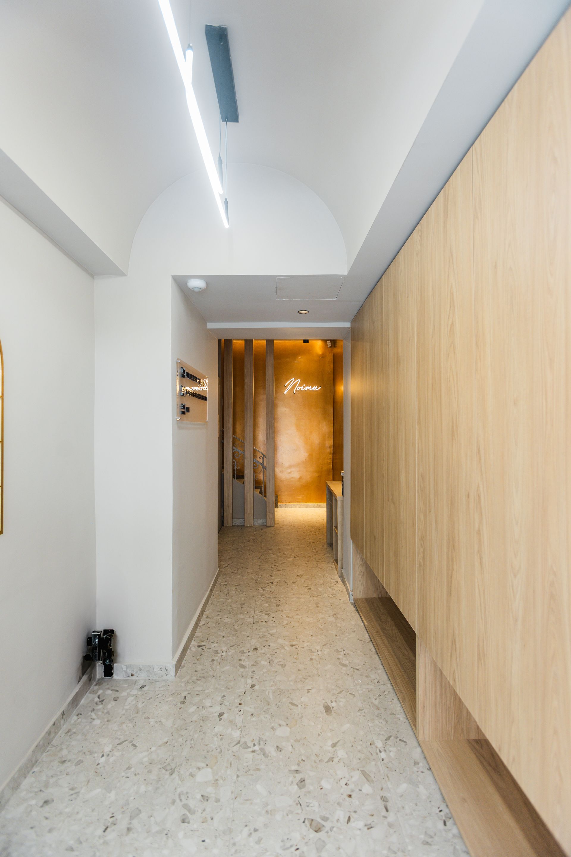 Narrow hallway with light wood paneling, terrazzo floor, and arched ceiling; bright lighting.