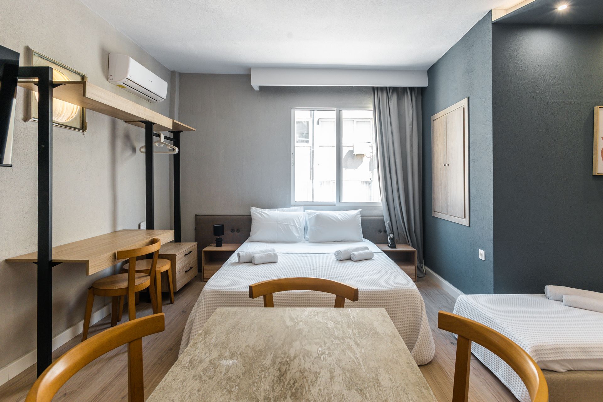 Hotel room with a bed, desk, table, and single bed; gray walls and neutral tones.
