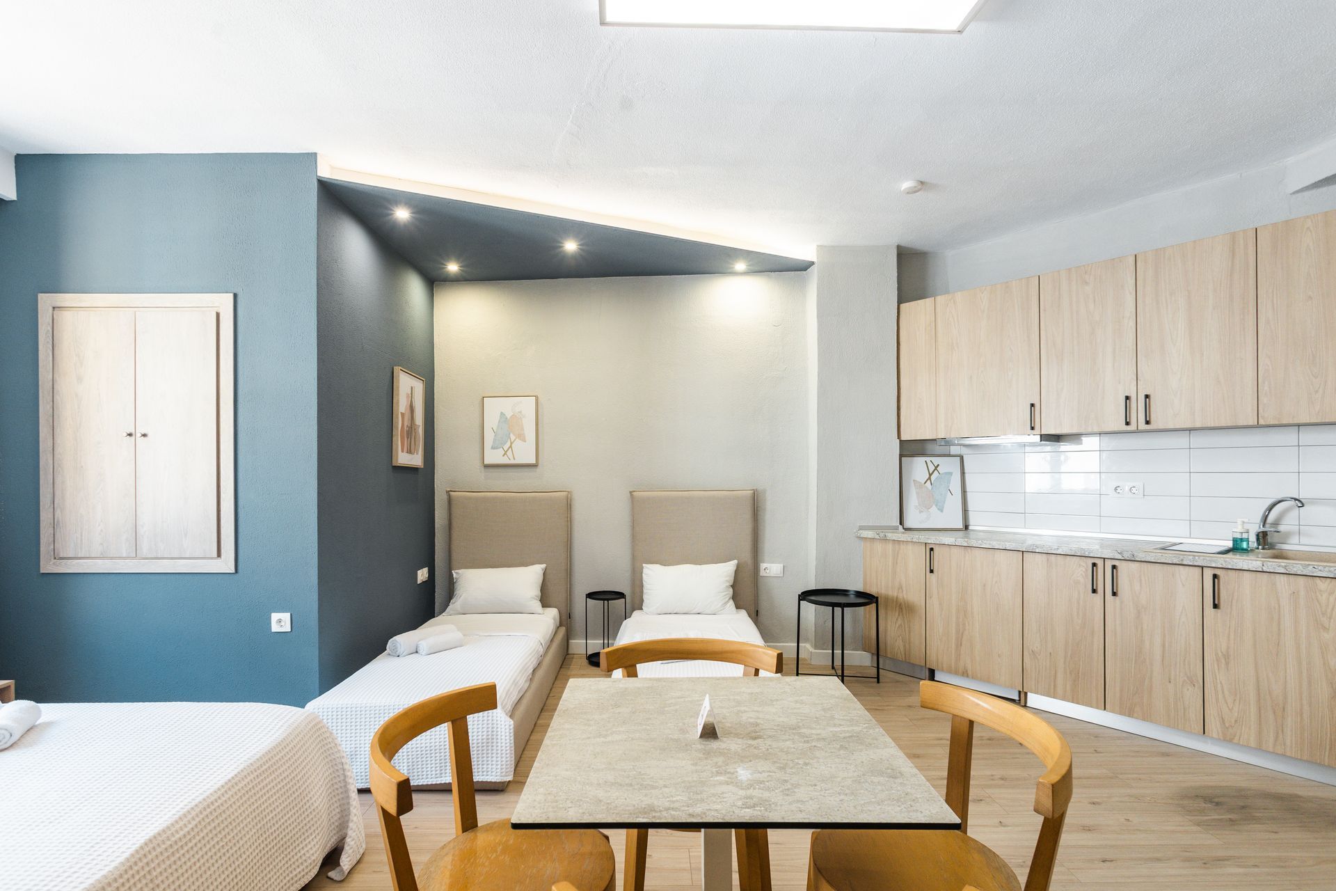 Studio apartment with beds, table, and kitchenette. Blue, gray, and light wood color scheme.