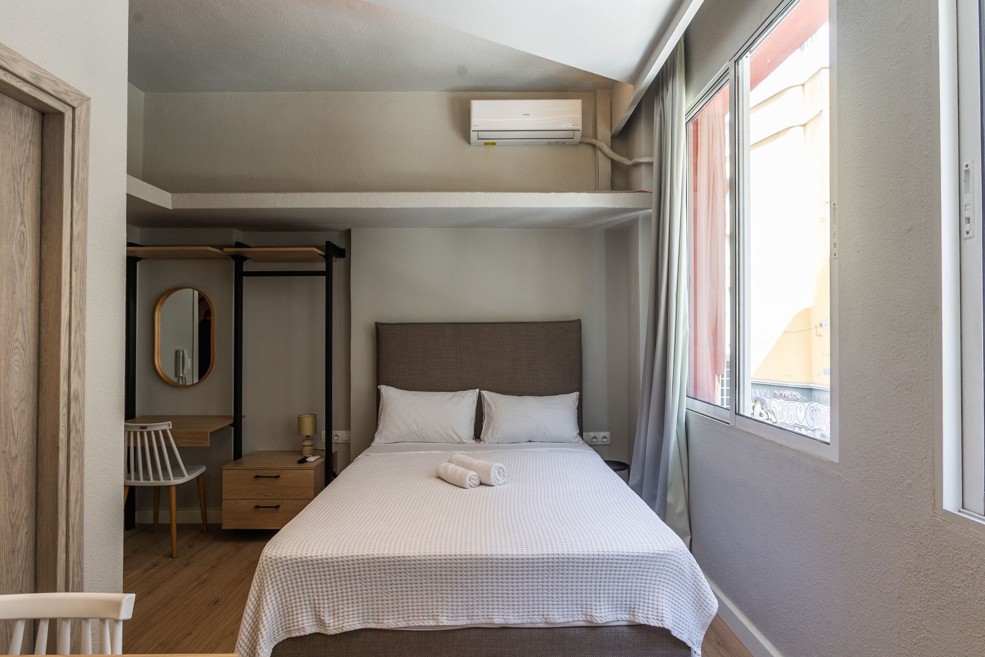 Bedroom with bed, desk, and windows. Light-colored walls and wood flooring.