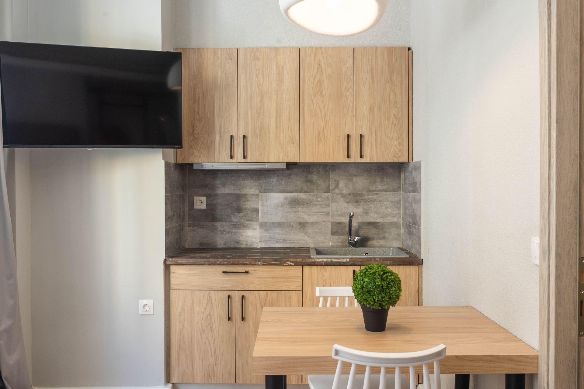 Small kitchen with wooden cabinets, countertop, and dining table. A TV is mounted on the wall.