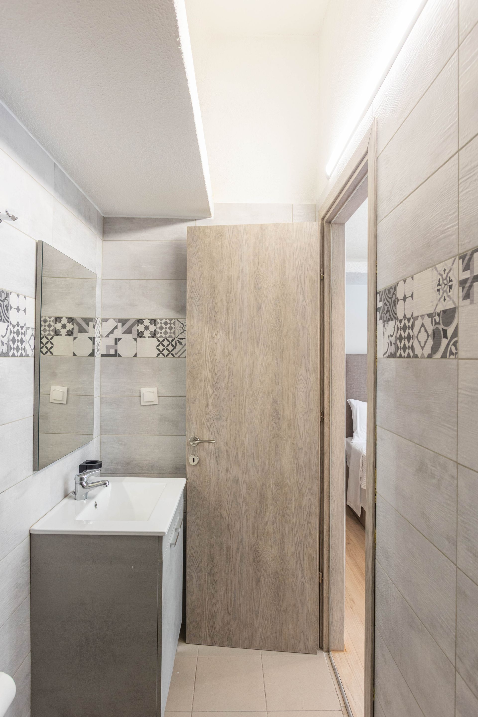 Bathroom with light gray tiled walls, vanity, open door to bedroom.