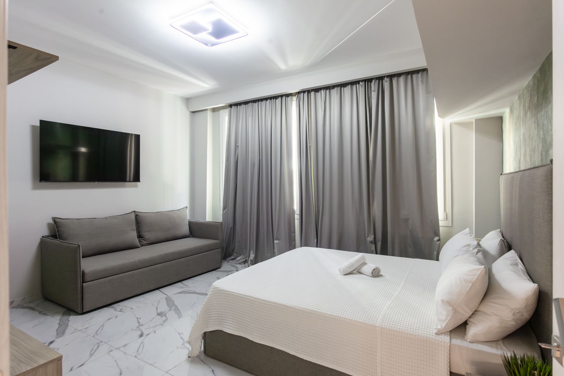 Bedroom with bed, sofa, TV, and gray curtains. Marble floors and white walls.