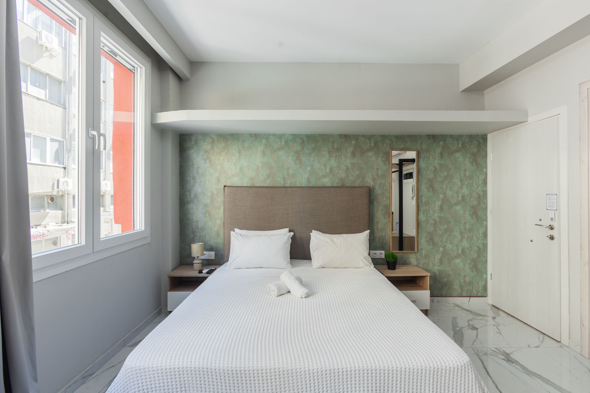 Bedroom with a bed, two nightstands, a window, and a door; light and airy design with marble floors.