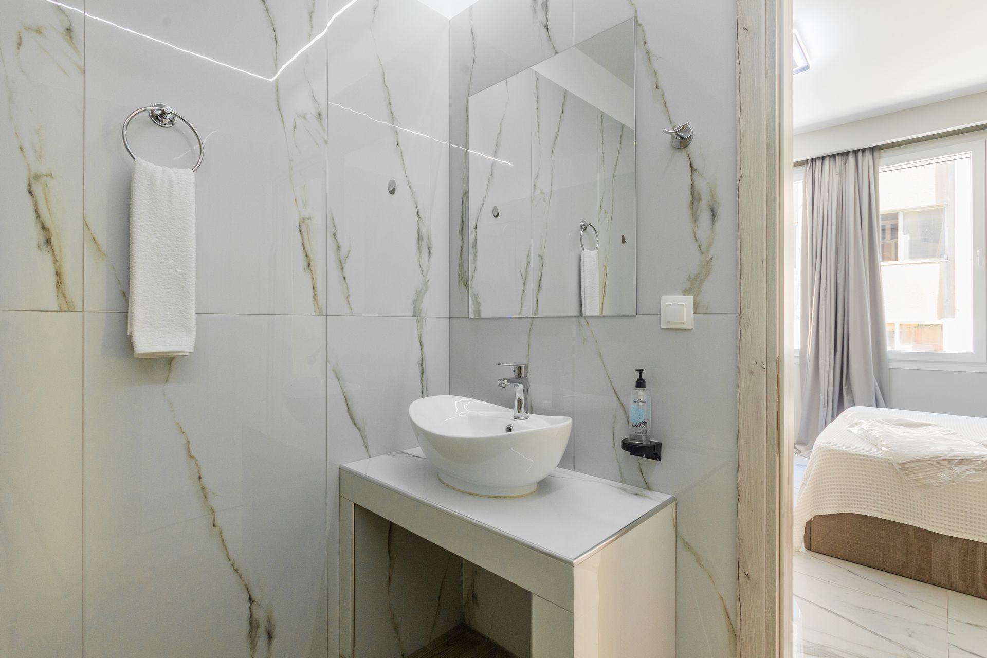 Modern bathroom with white marble-look tiles, a vessel sink, and an open doorway to a bedroom.