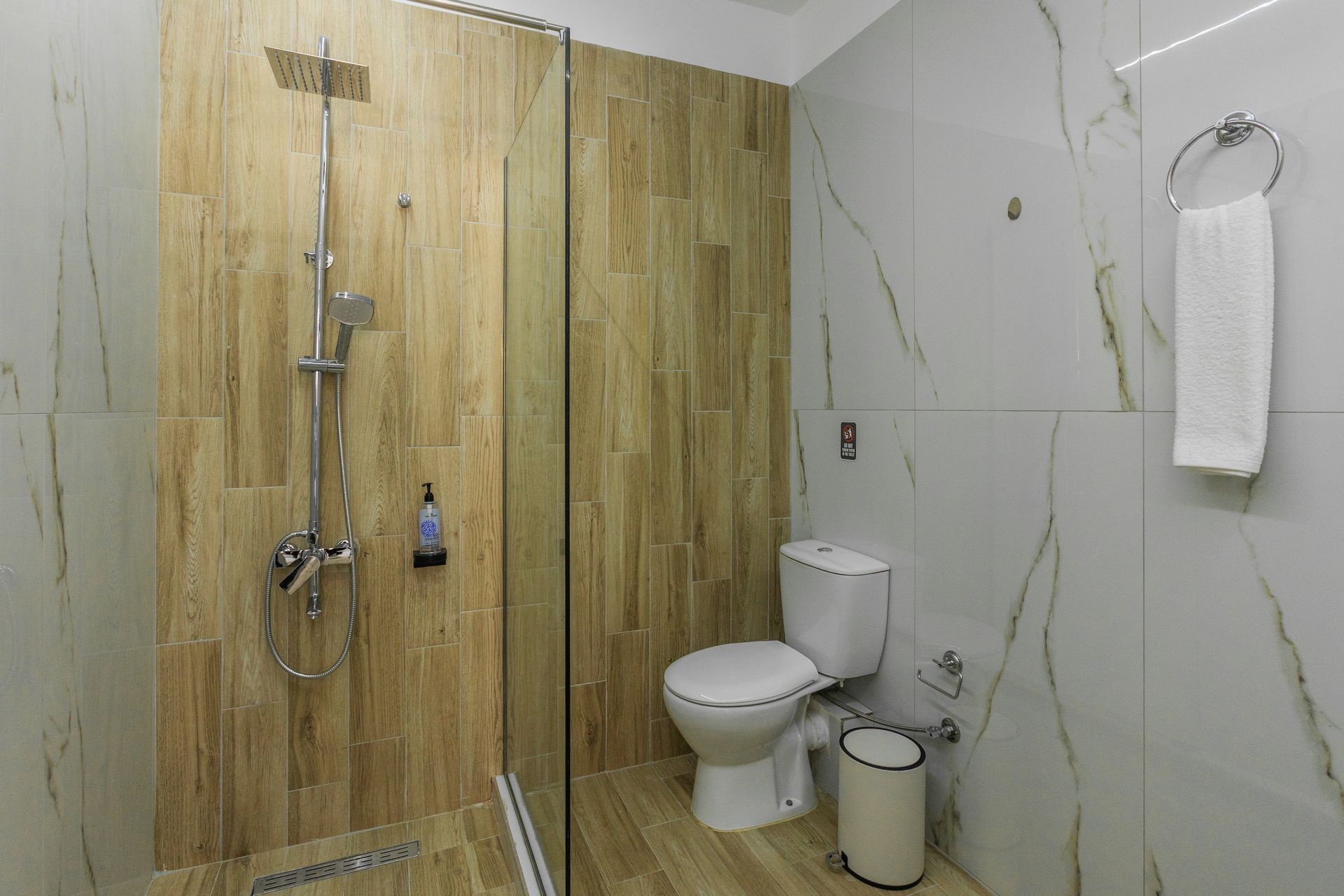 Bathroom with glass shower, toilet, towel, and beige and white marble-like tiles.