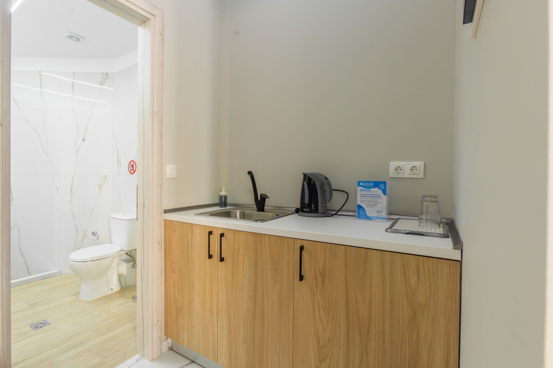 Small kitchenette with sink, kettle, and cabinets, next to an open doorway to a bathroom with a toilet.