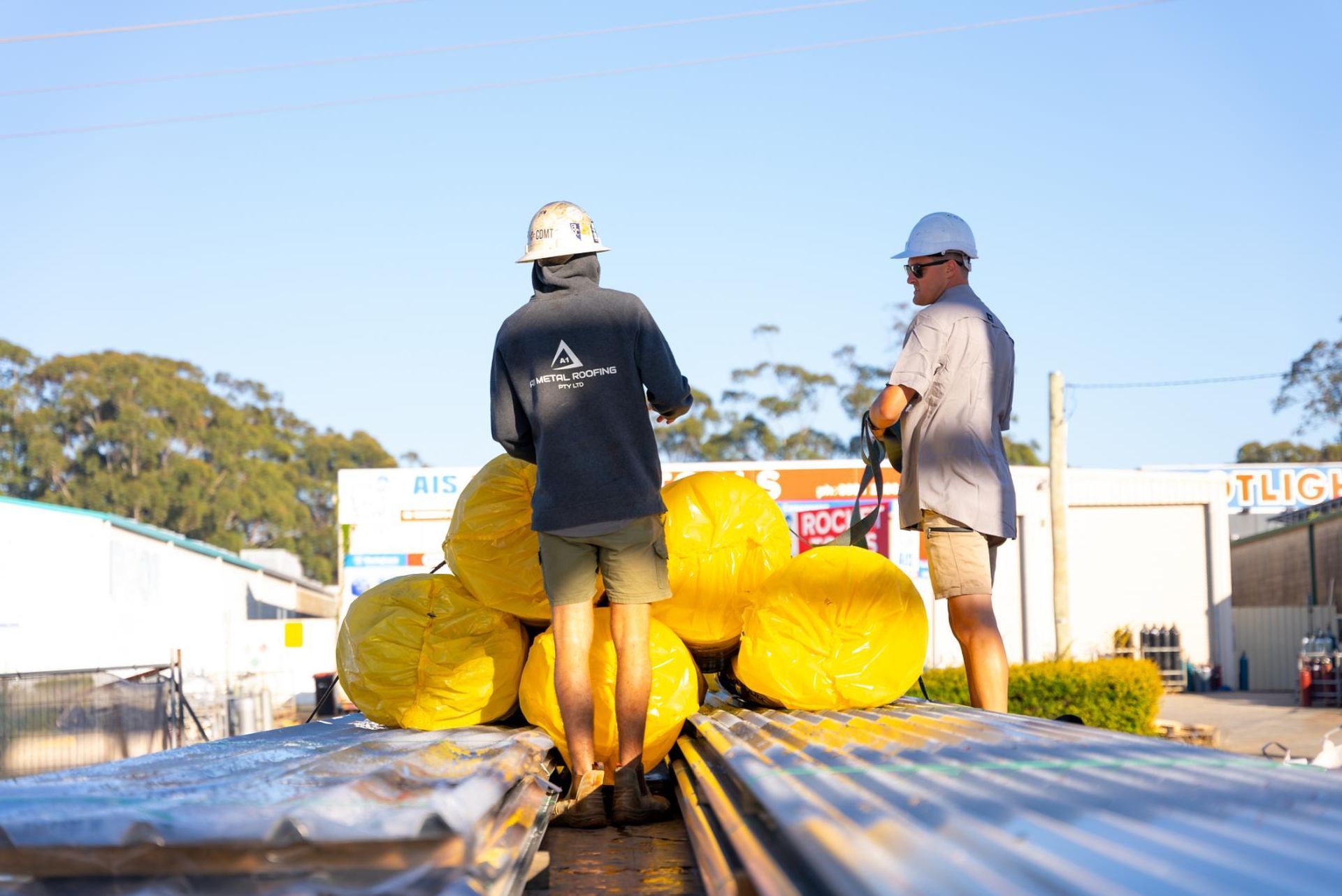 Two Men Are Standing Next to a Stack of Yellow Rolls of Insulation — Roofing Contractor in Wauchope, NSW