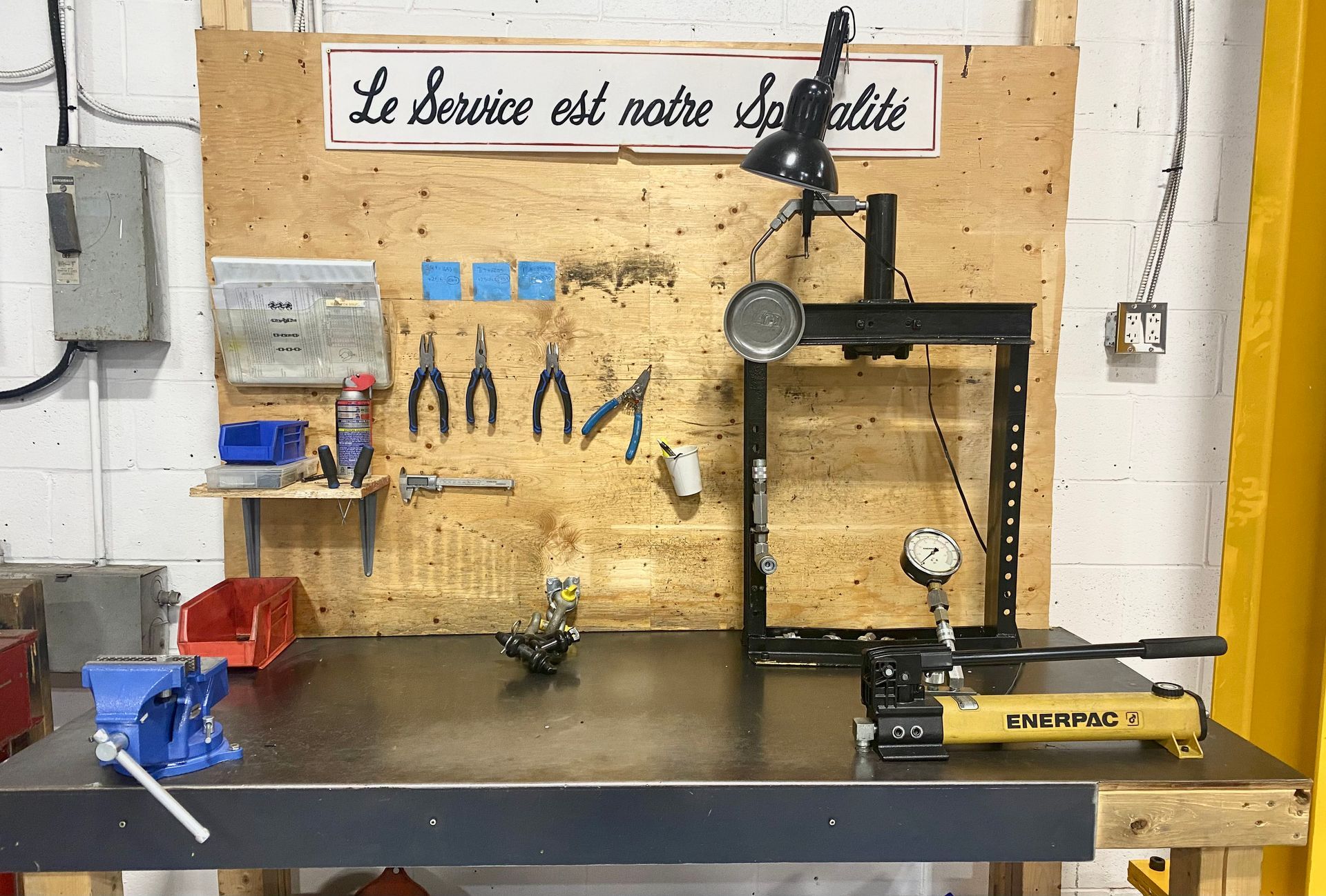 Établi d'atelier avec outils et équipements, comprenant une presse hydraulique, un étau et des outils suspendus.