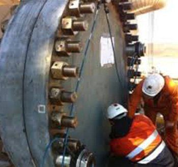 Des ouvriers portant des équipements de sécurité travaillent sur une grande structure industrielle, probablement une cuve ou une canalisation, fixée par des boulons.