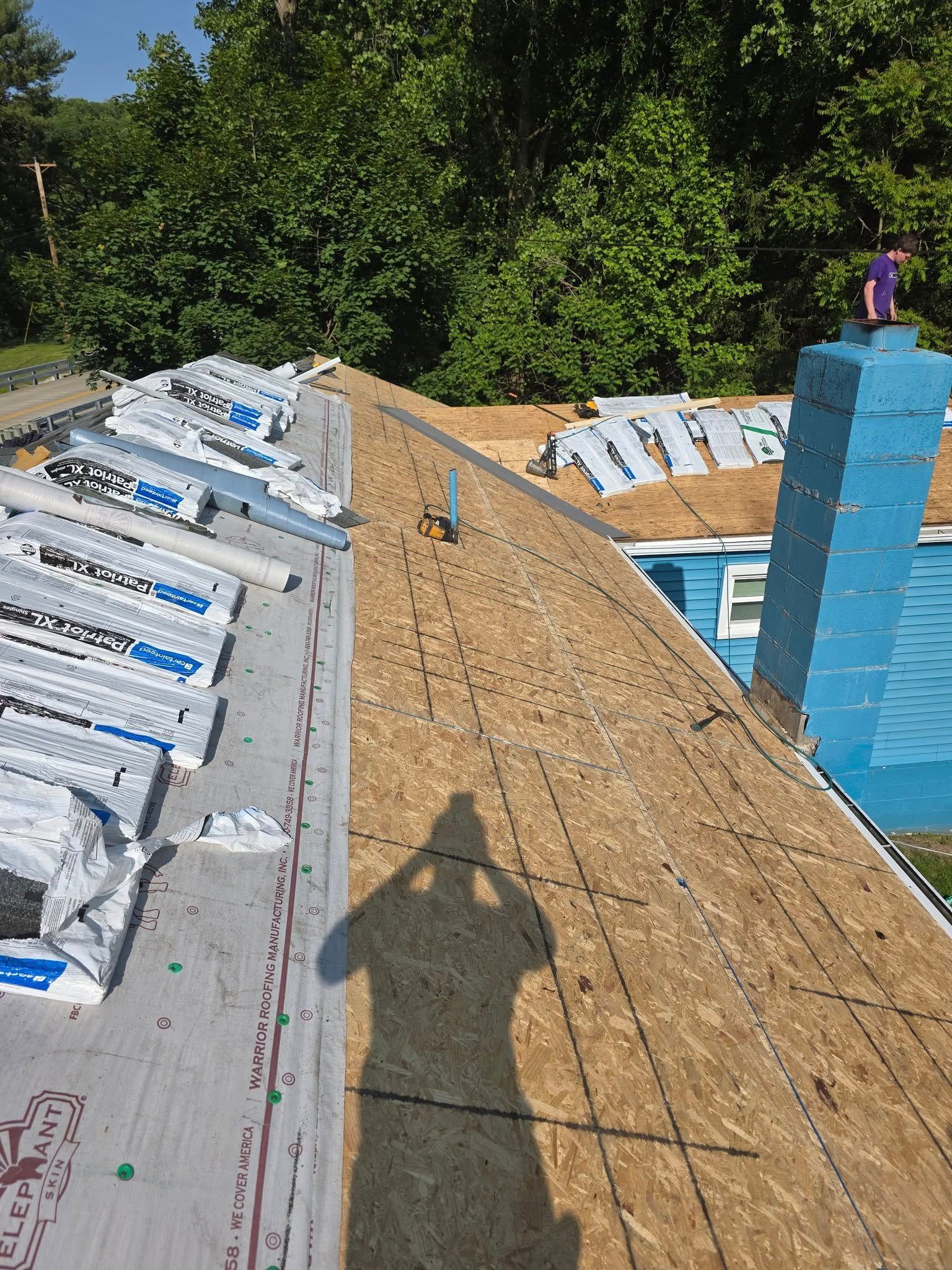Roofing construction with plywood, tar paper, shingles, and a blue chimney. Shadow of a person taking the photo.