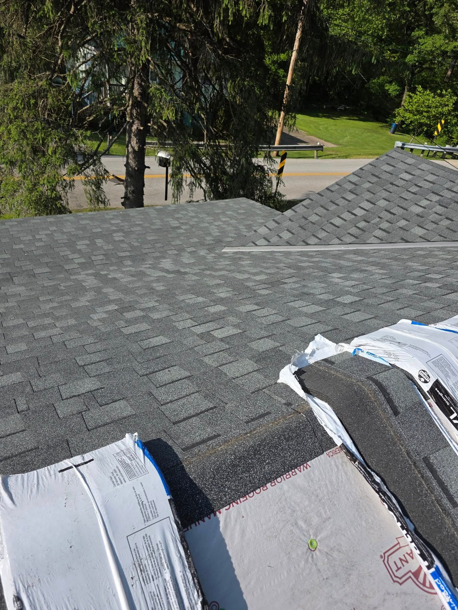 View of a dark gray shingled roof with materials, trees, road, and houses in the background on a sunny day.