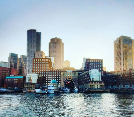 The Boston skyline viewed from the harbor, featuring tall buildings and boats docked at a waterfront pier at sunset.