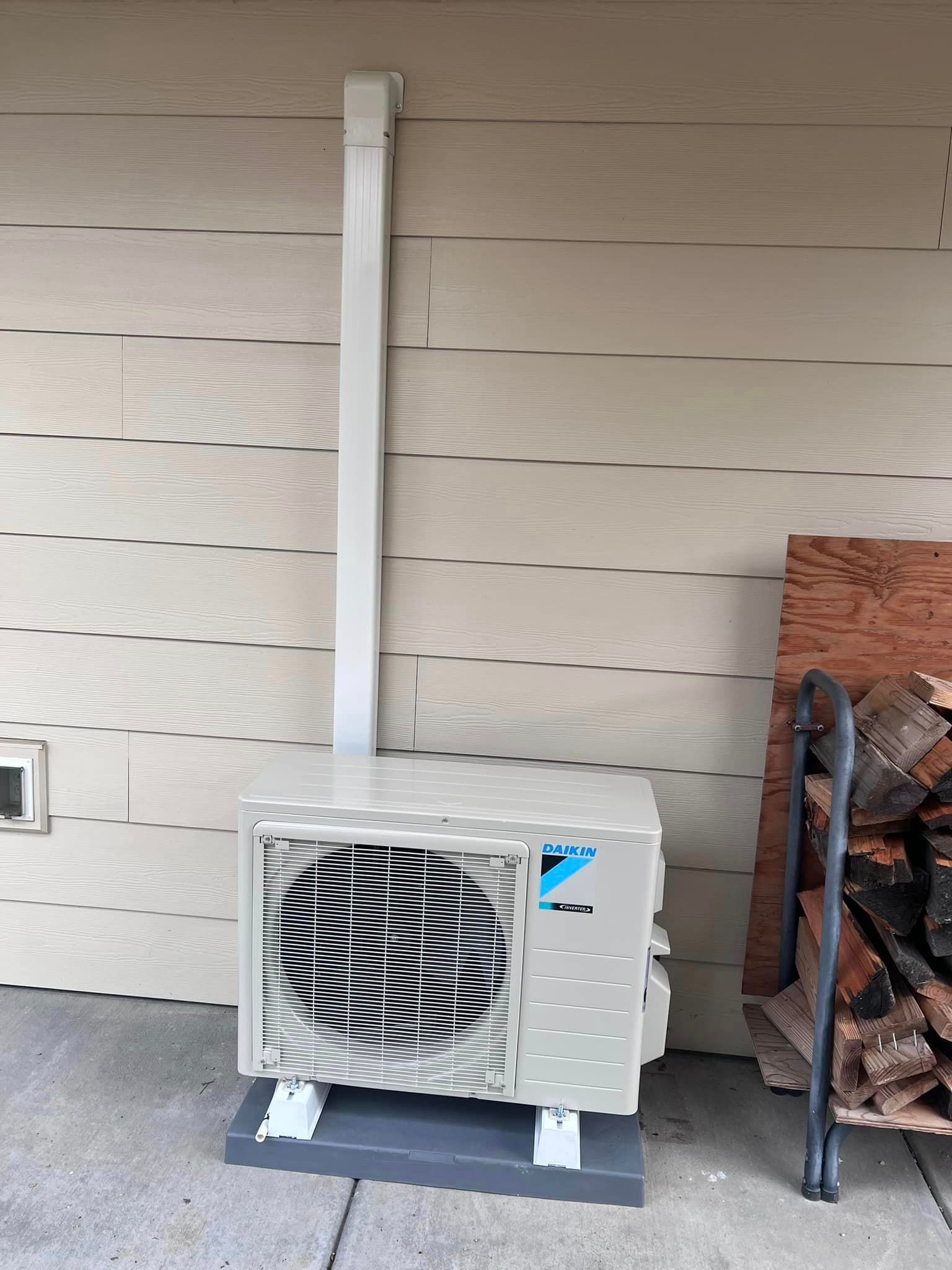 A white air conditioner is sitting on the side of a building next to a pile of logs.