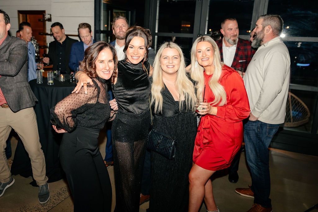 Four women smiling together at party with drinks while other guests mingle in background.