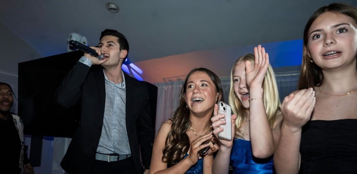 A group of young people are watching a man speak into a microphone at a party.