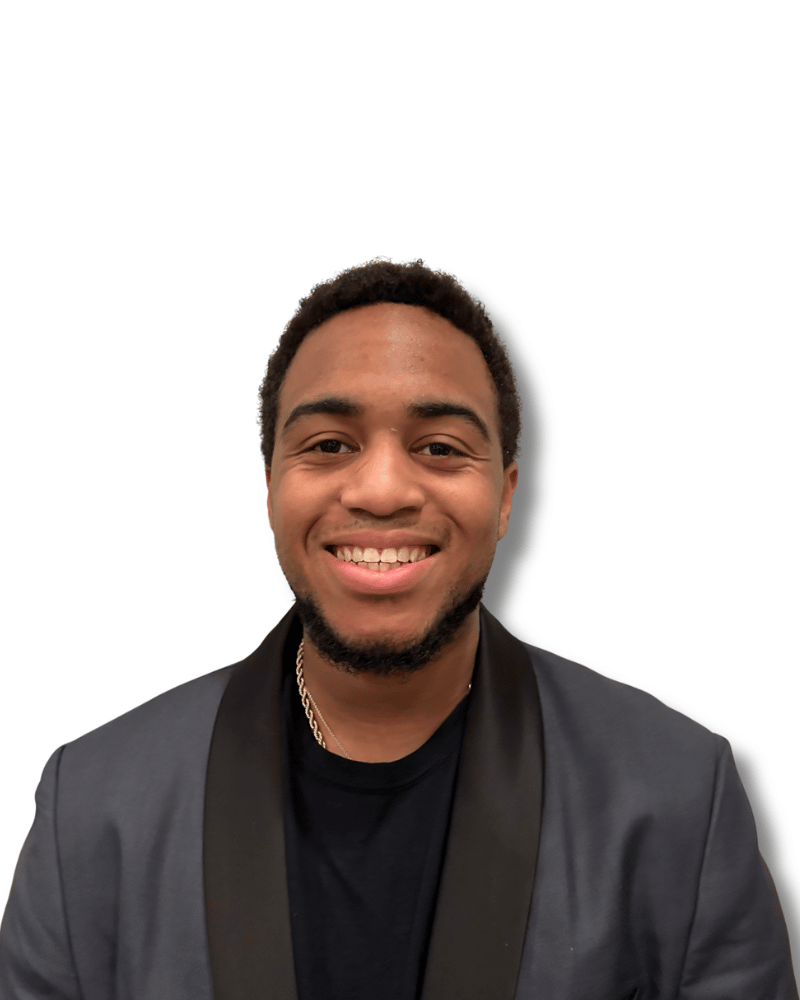 A person with a beard smiling while wearing a dark grey blazer over a black shirt against a white background.